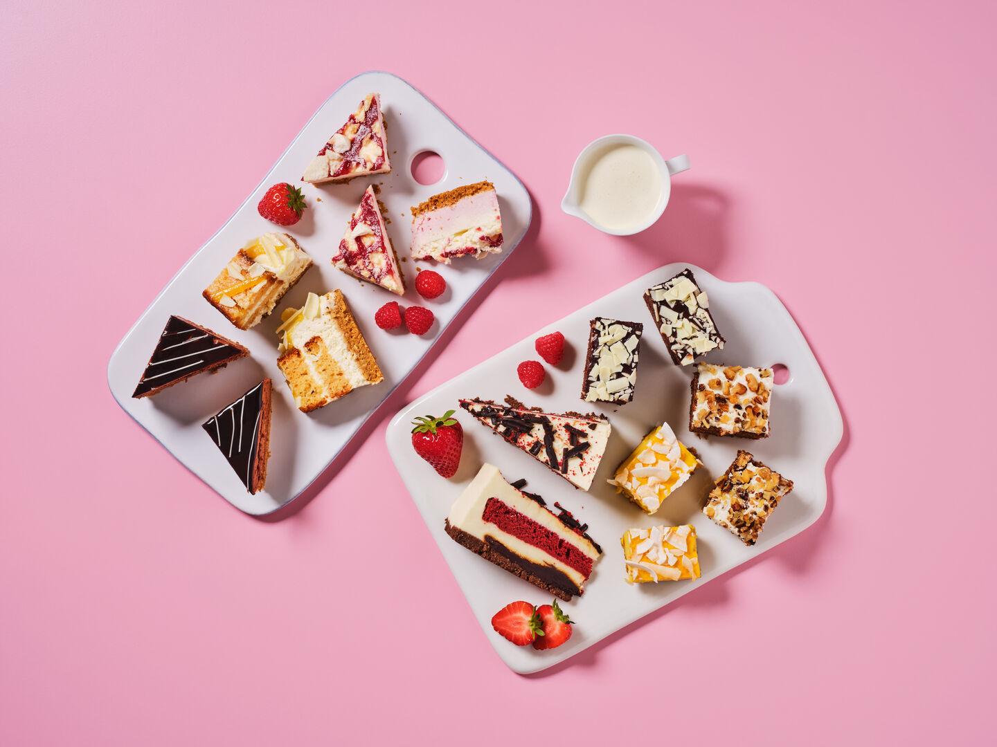 A selection of cake slices and traybakes, including chocolate, red velvet, lemon and fruit sponges, garnished with strawberries and raspberries, with a jug of cream, on a pink background.