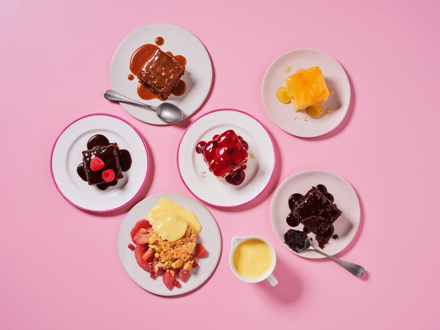 A selection of hot puddings on a pink background, including sticky toffee pudding, chocolate sponge with sauce and berries, treacle sponge, and fruit crumble with custard.