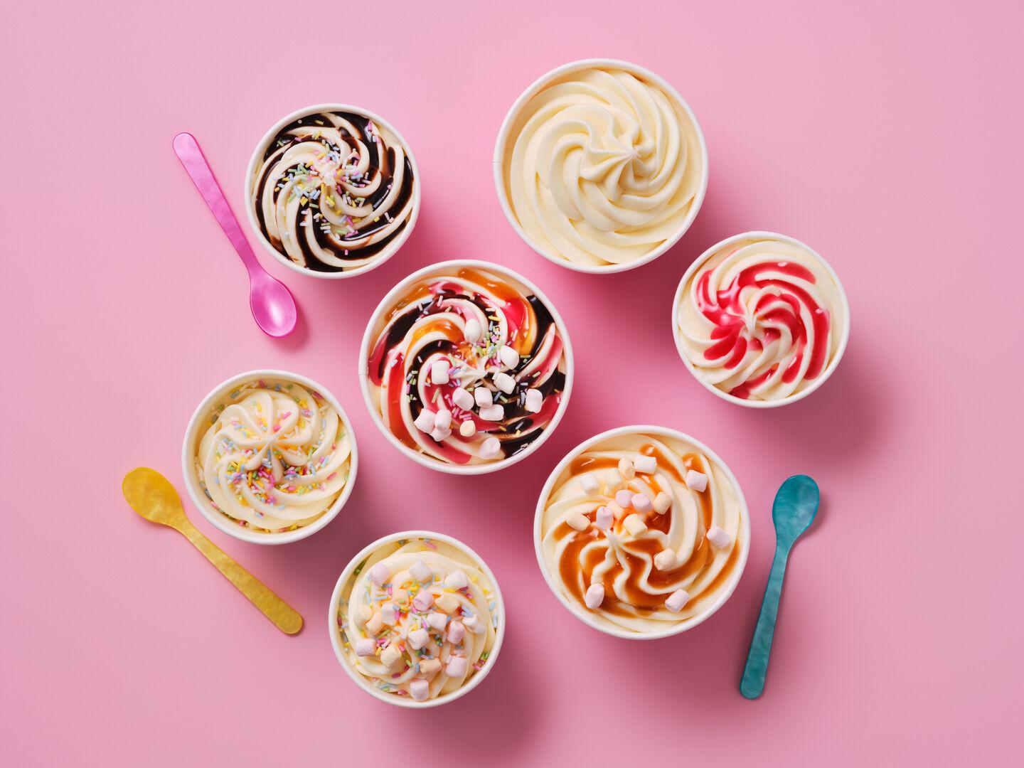 Cups of soft-serve ice cream with toppings including sprinkles, marshmallows, chocolate, caramel and strawberry sauce, with colourful spoons, on a pink background.