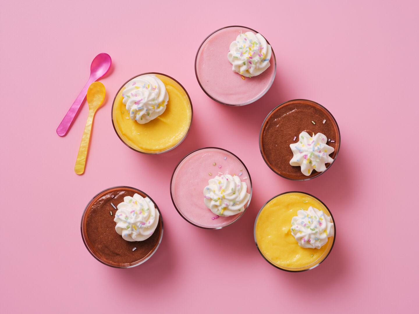 Pots of banana, strawberry and chocolate mousse topped with whipped cream and sprinkles, with colourful spoons, on a pink background.