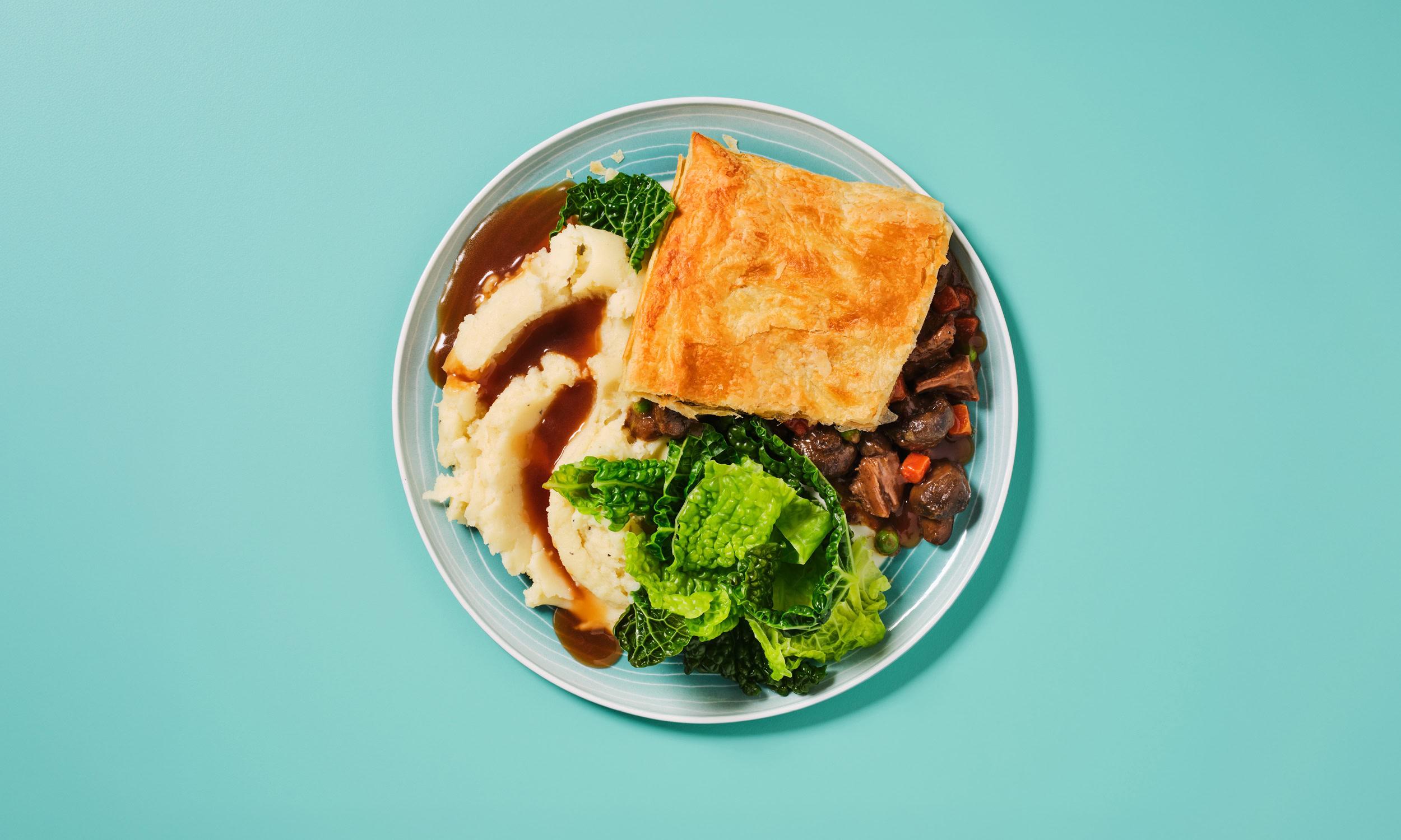 A overhead shot of a plate of beef and mushroom pie with golden puff pastry, served with creamy mashed potato topped with rich gravy, and steamed savoy cabbage and kale, on a teal background.