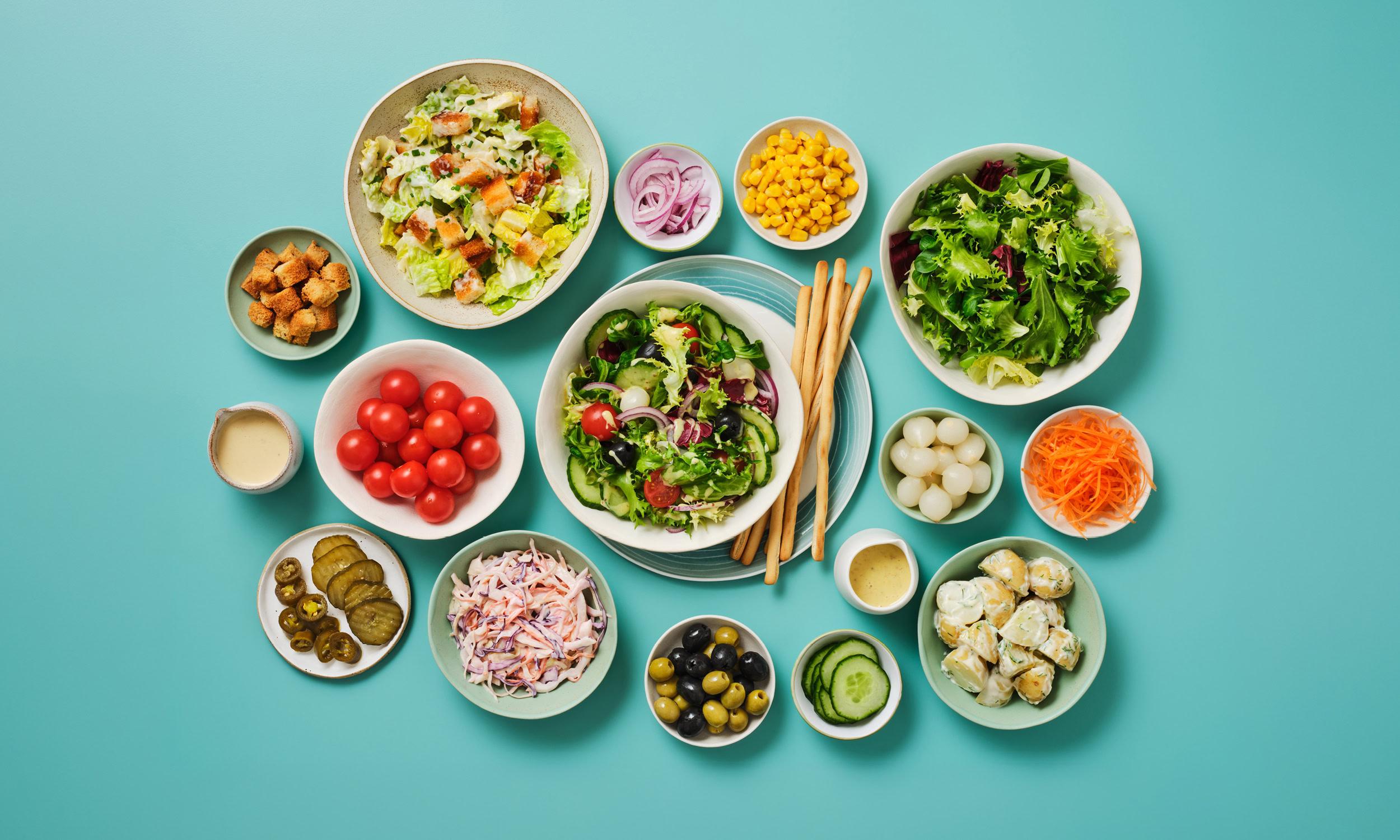 A flatlay of salad bar dishes and toppings arranged on a teal background. A central mixed leaf salad with tomato, cucumber, olive, and red onion is served on a plate with breadsticks. Surrounding bowls and dishes include a Caesar salad with croutons, a mixed green leaf salad, coleslaw, cherry tomatoes, pickled gherkins and jalapeños, sweetcorn, sliced red onion, grated carrot, mixed green and black olives, cucumber slices, pickled silverskin onions, herbed potato salad, and two small pots of dressing.