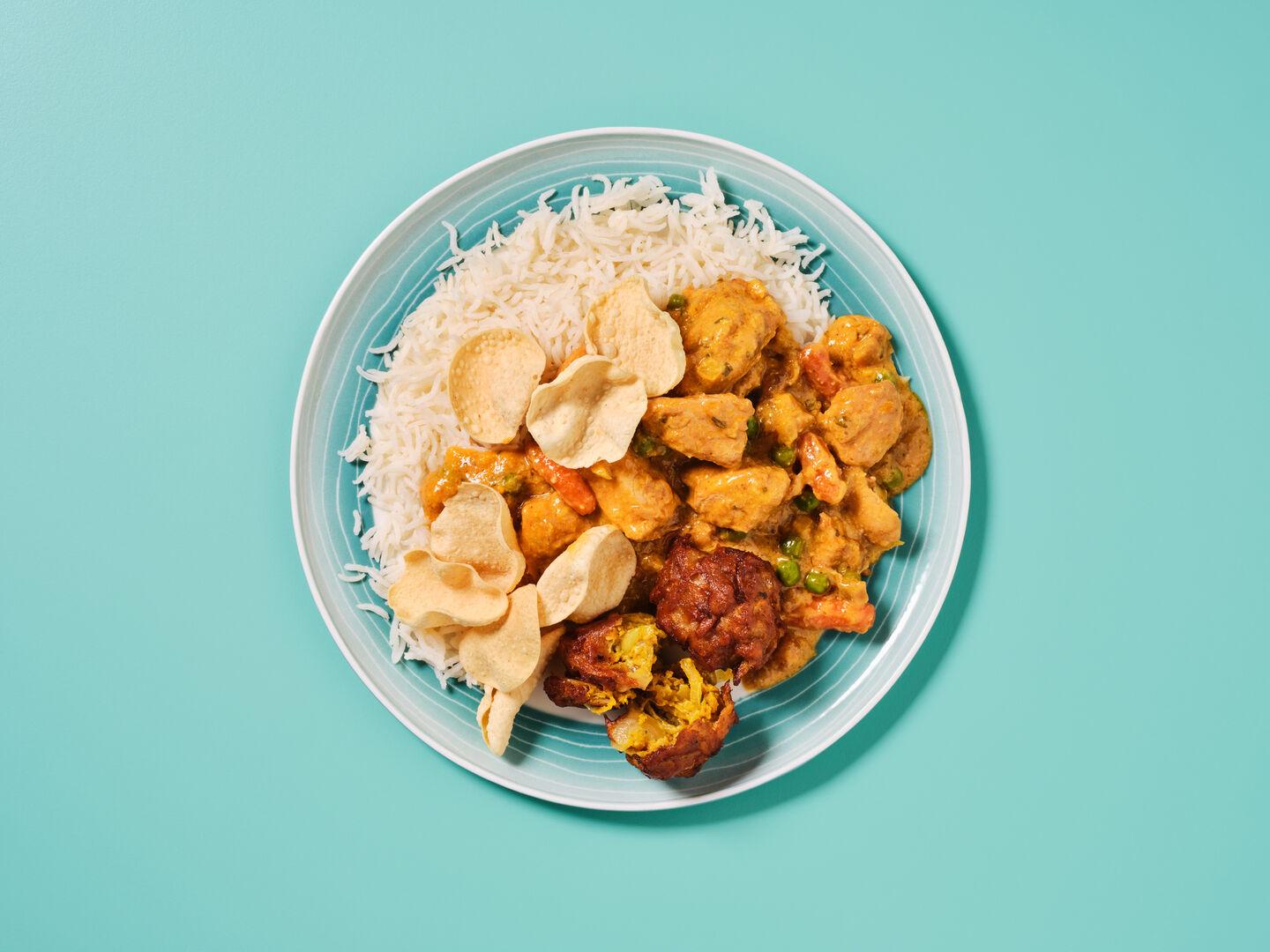 A plate of chicken curry with peas and carrots, served with rice, poppadoms and a vegetable pakora, on a teal background.