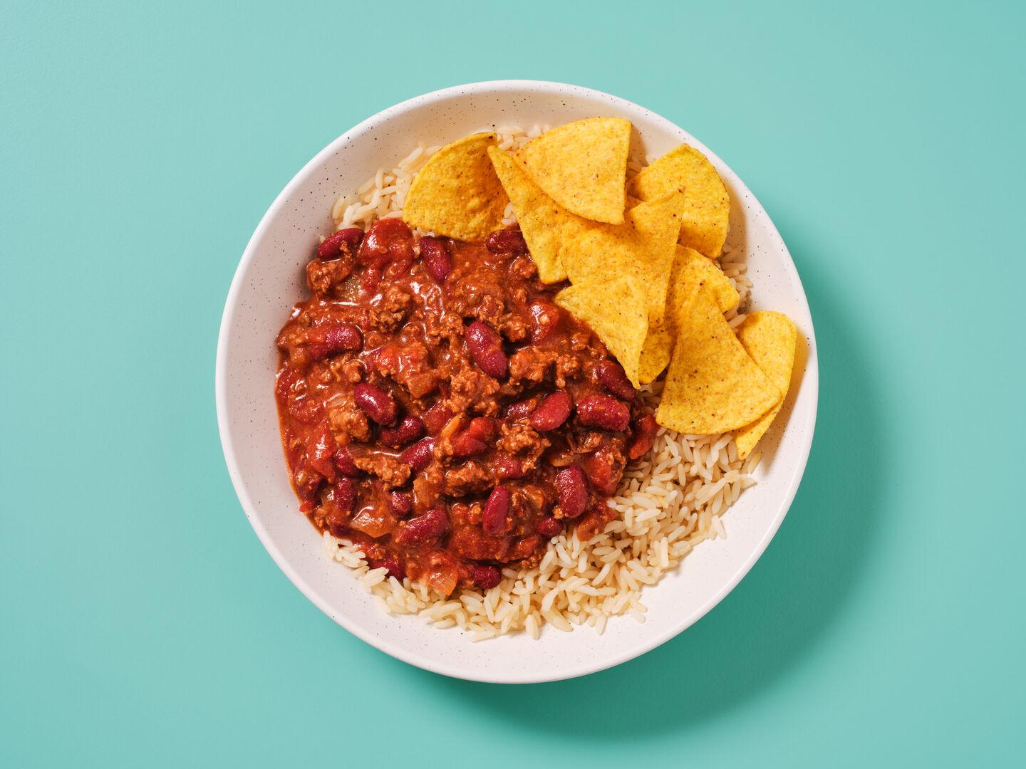 A bowl of chilli con carne with kidney beans, served on rice with tortilla chips, on a teal background.