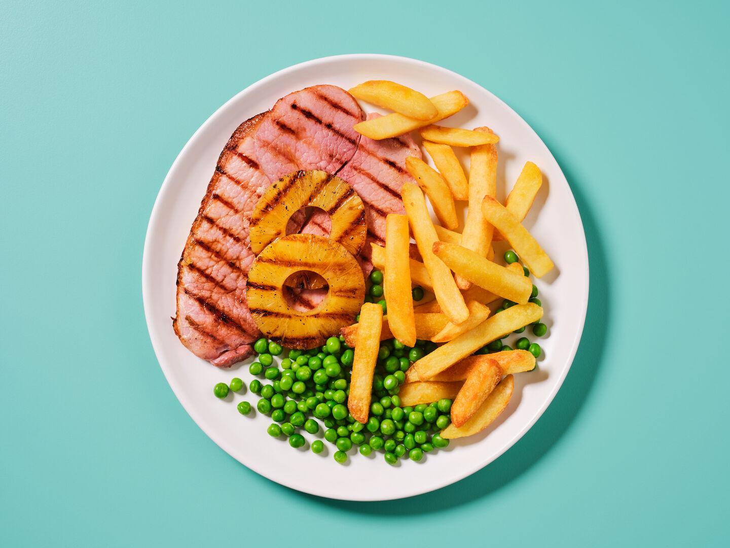 A plate of grilled gammon steak with pineapple rings, served with chips and peas, on a teal background.