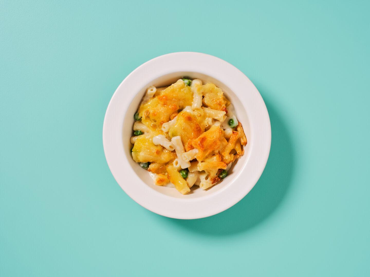 A bowl of baked macaroni cheese with peas, on a teal background.