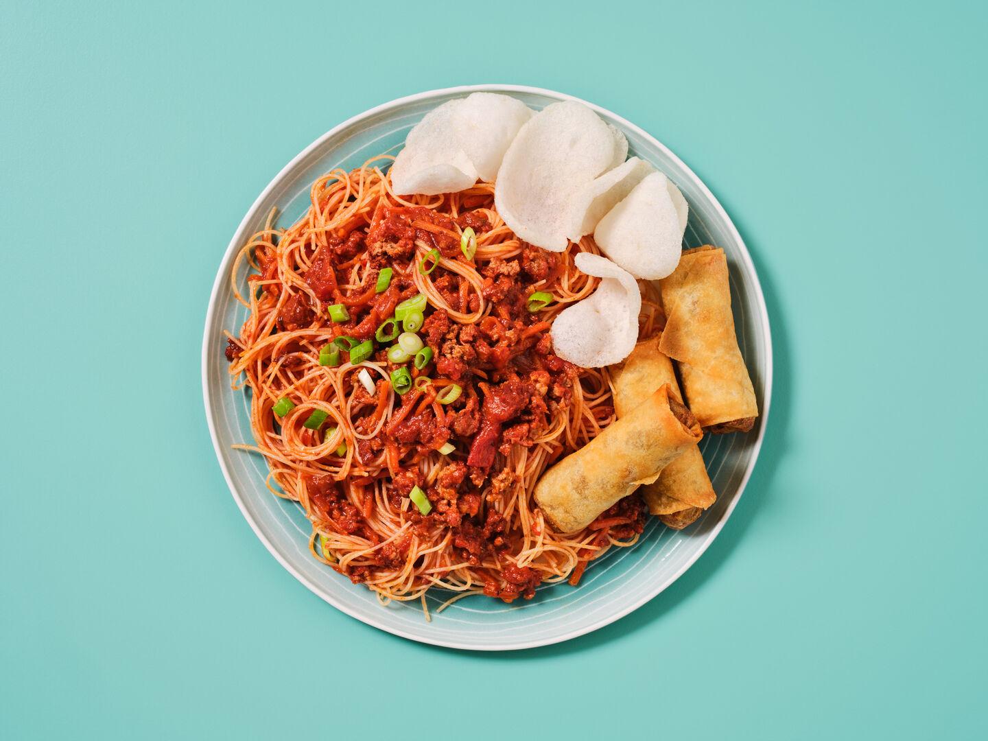 A plate of noodles with a spiced sauce, garnished with spring onion, served with spring rolls and prawn crackers, on a teal background.