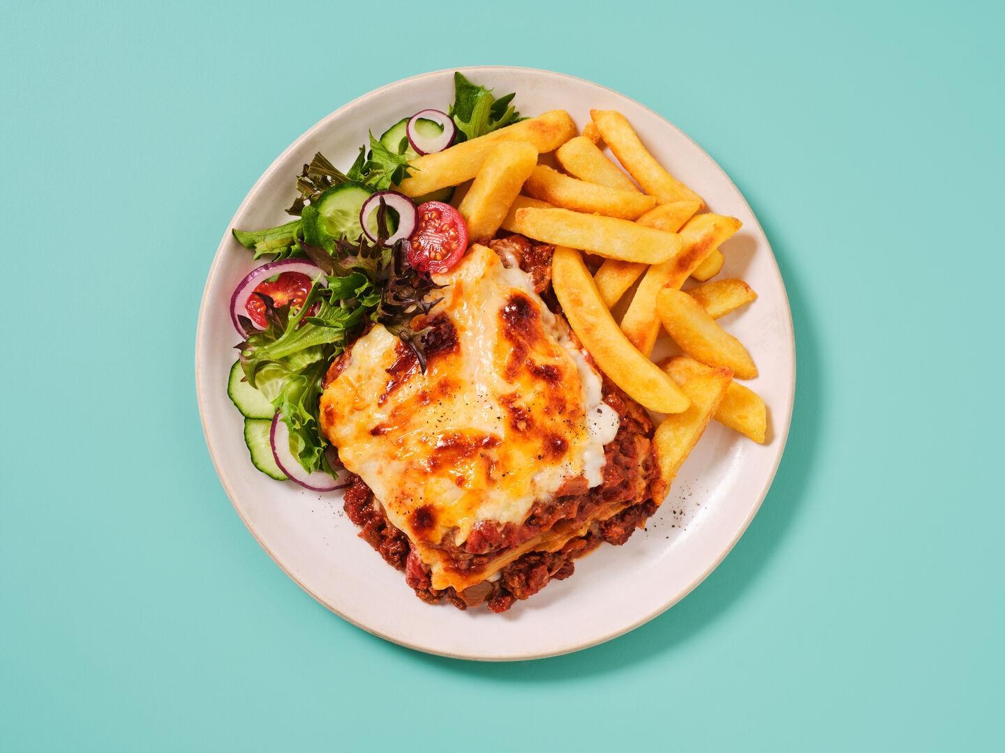 A plate of lasagne served with chips and a side salad, on a teal background.