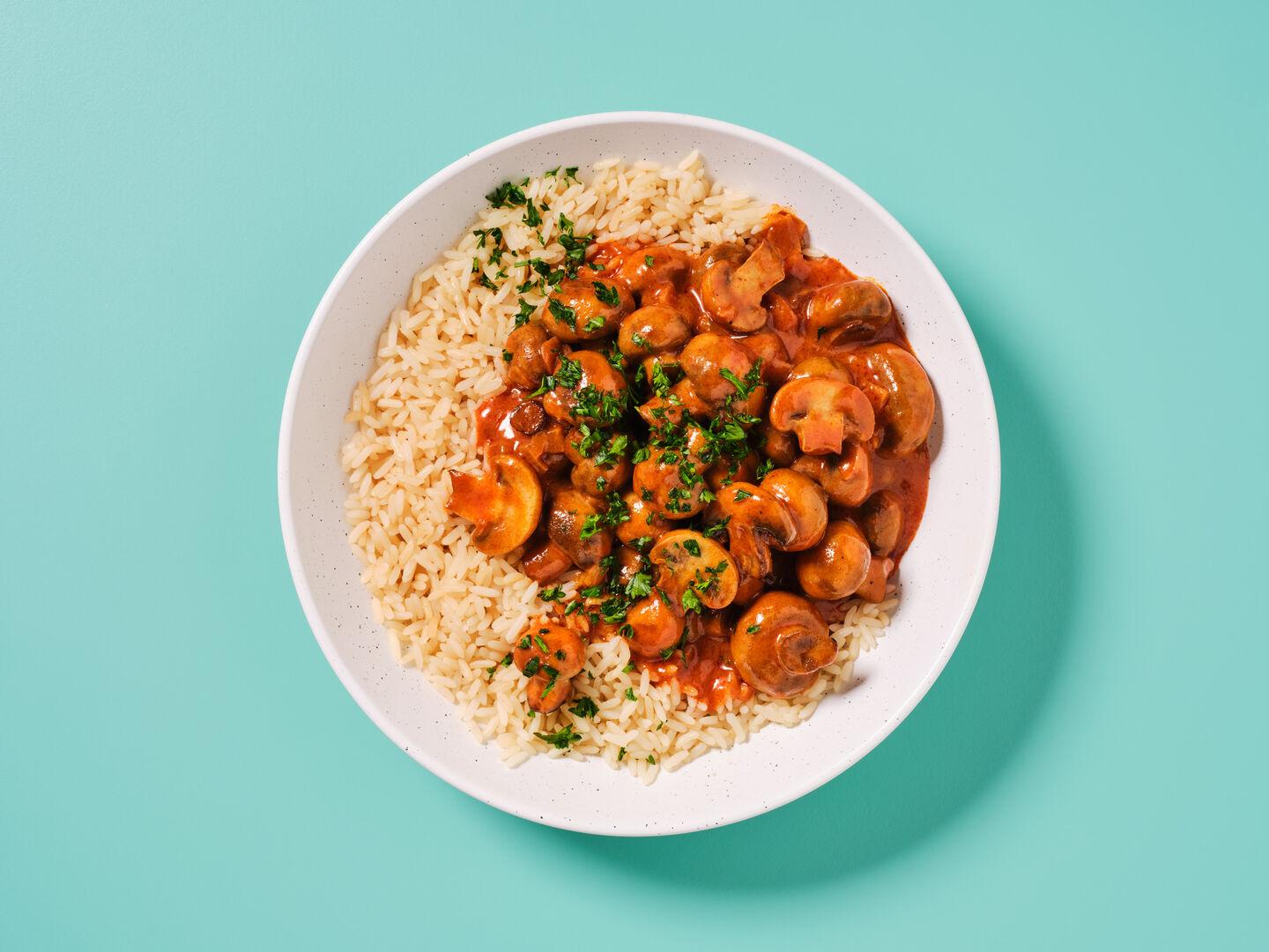A plate of mushrooms in a tomato sauce garnished with parsley, served on rice, on a teal background.