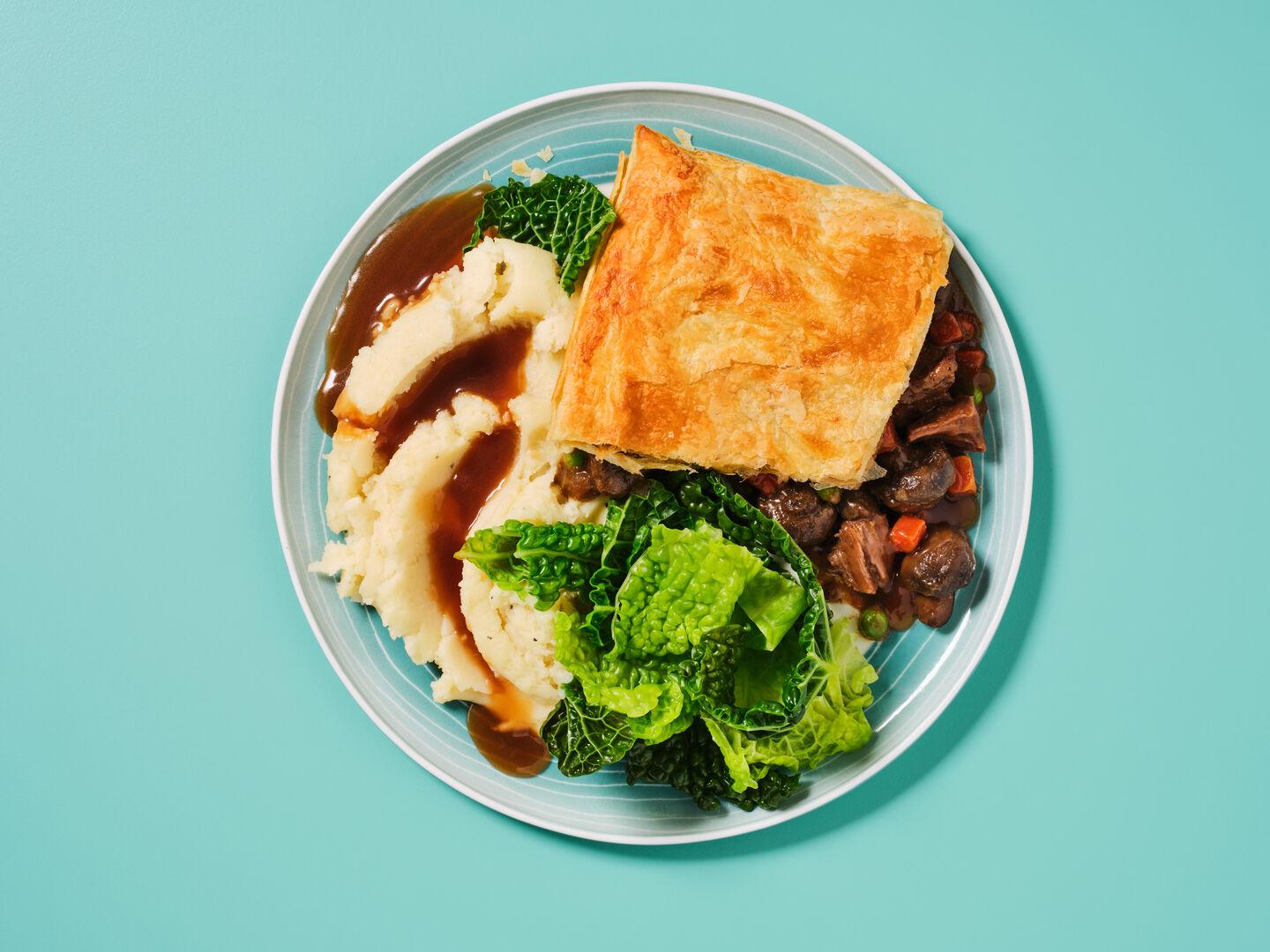 A plate of steak and ale pie with mashed potato, savoy cabbage and gravy, on a teal background.