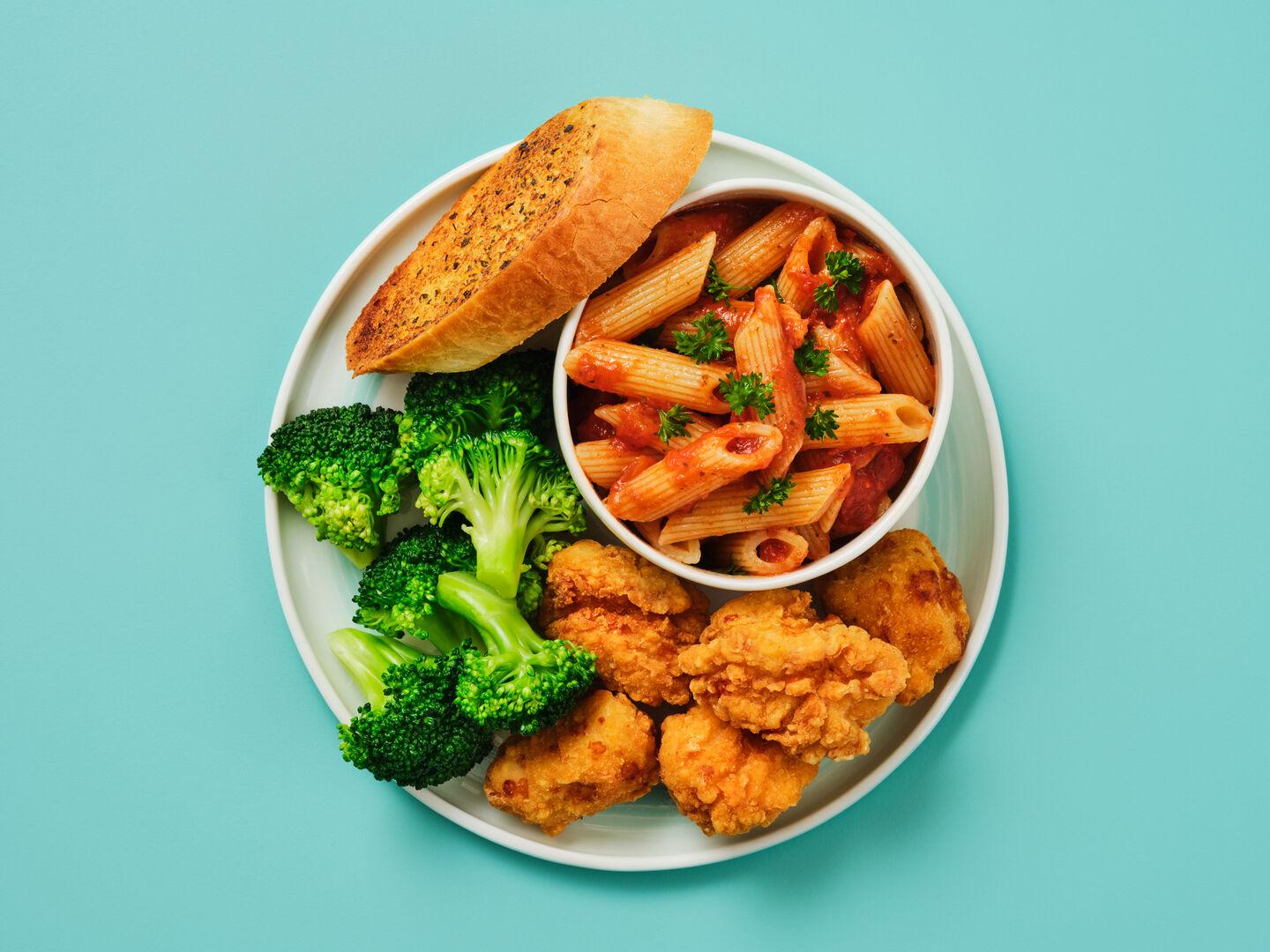 A plate of chicken goujons with broccoli, garlic bread and penne pasta in tomato sauce, on a teal background.