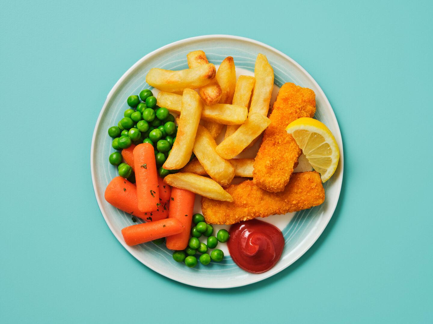 A plate of breaded fish fingers with chips, peas and carrots, served with a lemon wedge and ketchup, on a teal background.