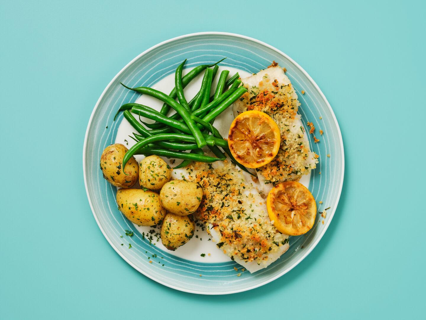 A plate of breadcrumb-crusted fish topped with grilled lemon, served with green beans and new potatoes, on a teal background.
