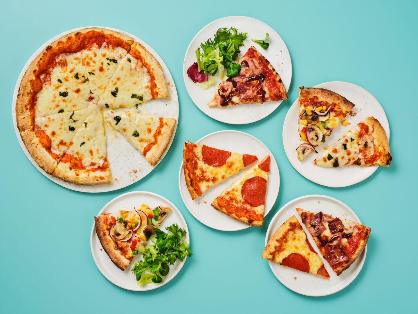 A whole margherita pizza alongside plates of pizza slices in various toppings, including pepperoni, BBQ bacon and vegetable, on a teal background.