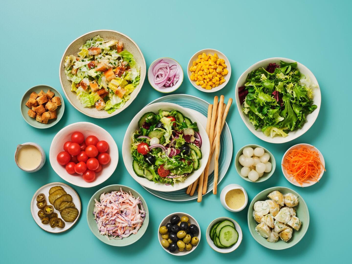 An array of salad bowls on a teal background, including Caesar salad, mixed leaf salad, coleslaw, potato salad, and side bowls of tomatoes, olives, sweetcorn, red onion, croutons and breadsticks.