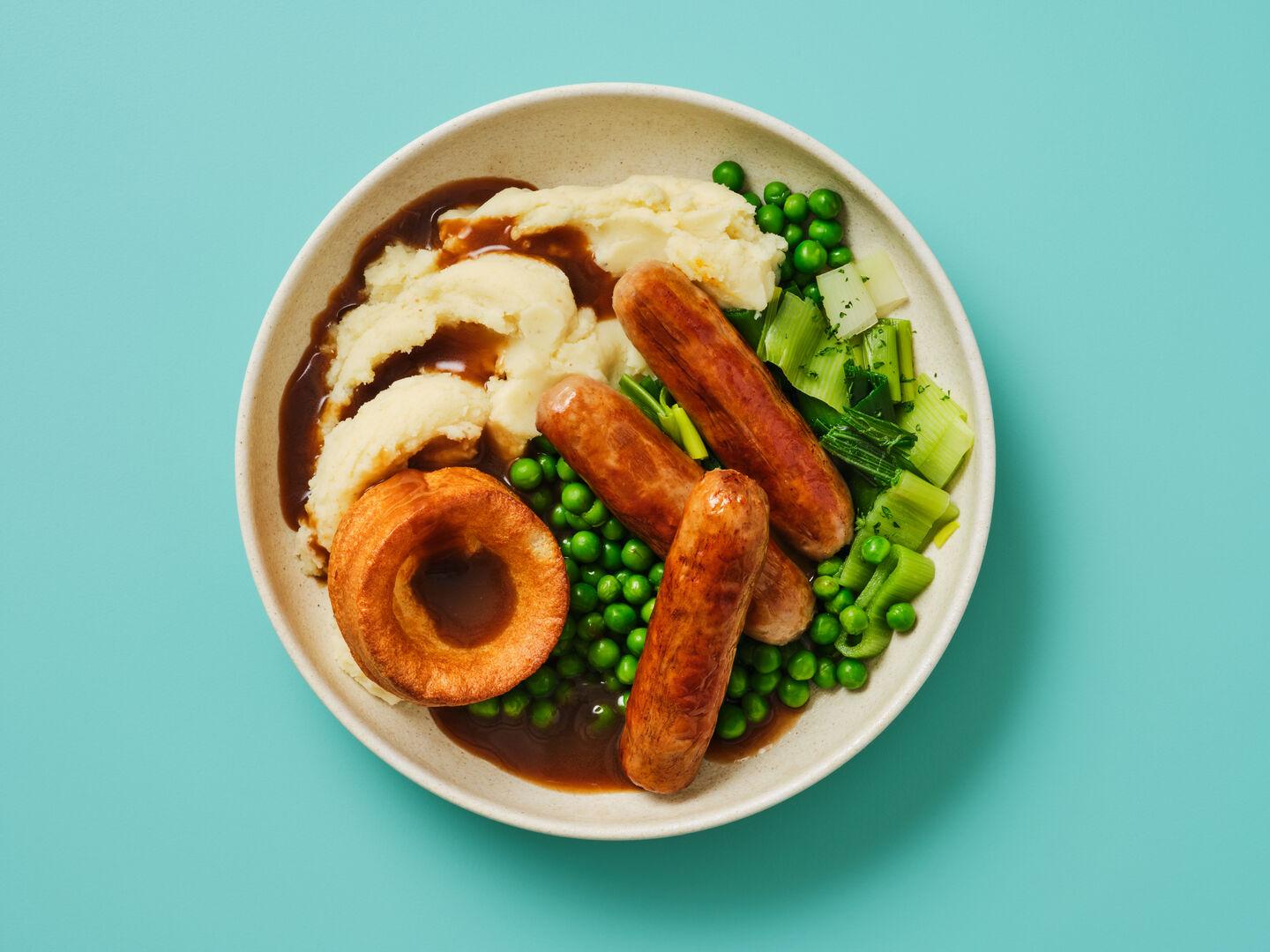 A plate of sausages with mashed potato, a Yorkshire pudding, peas, leeks and gravy, on a teal background.