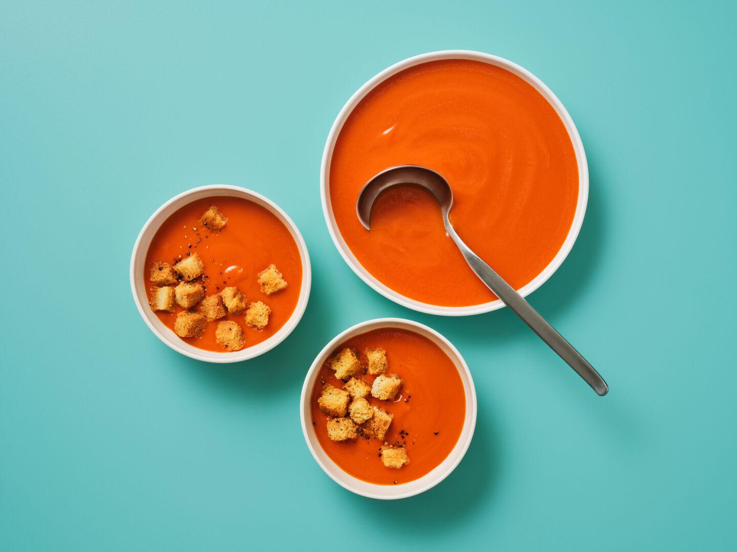 Three bowls of tomato soup topped with croutons, with a spoon, on a teal background.