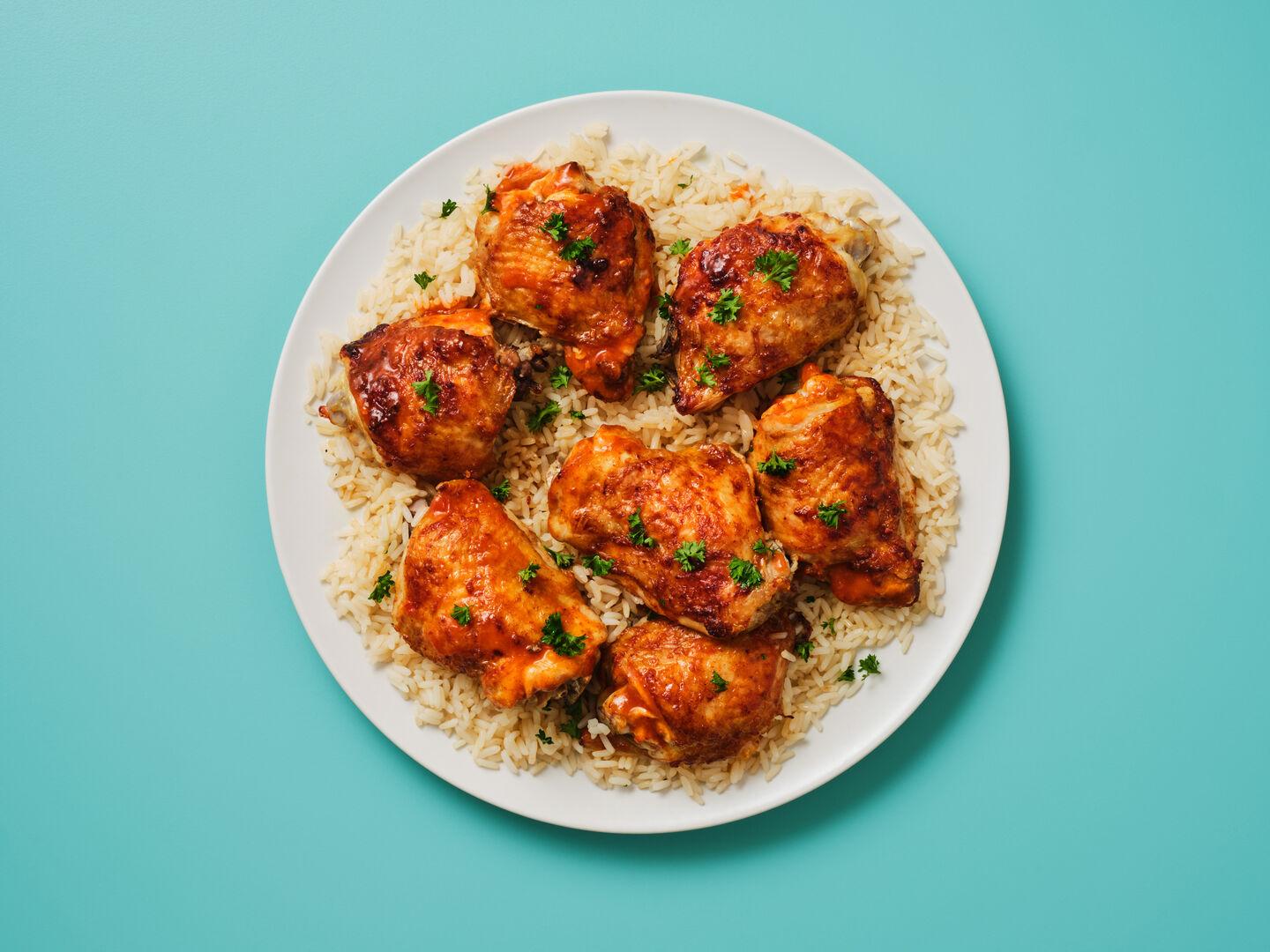 A plate of glazed chicken thighs garnished with parsley, served on rice, on a teal background.