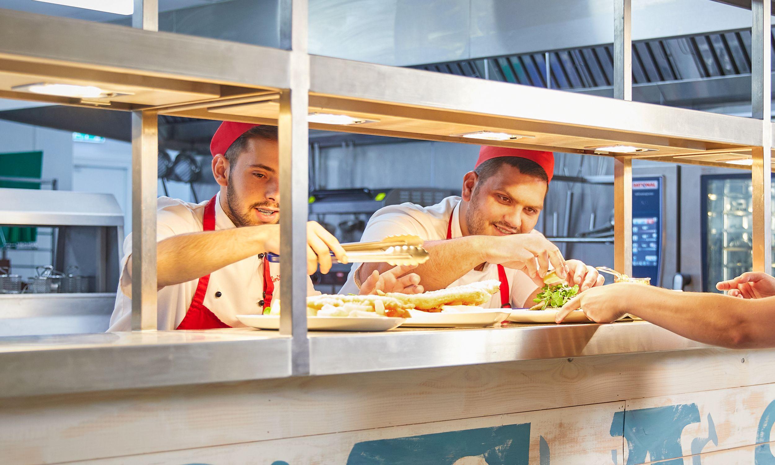 Two chefs in red caps and aprons plating up fish, chips, and salad at a live cooking station counter, handing a plate to a guest.