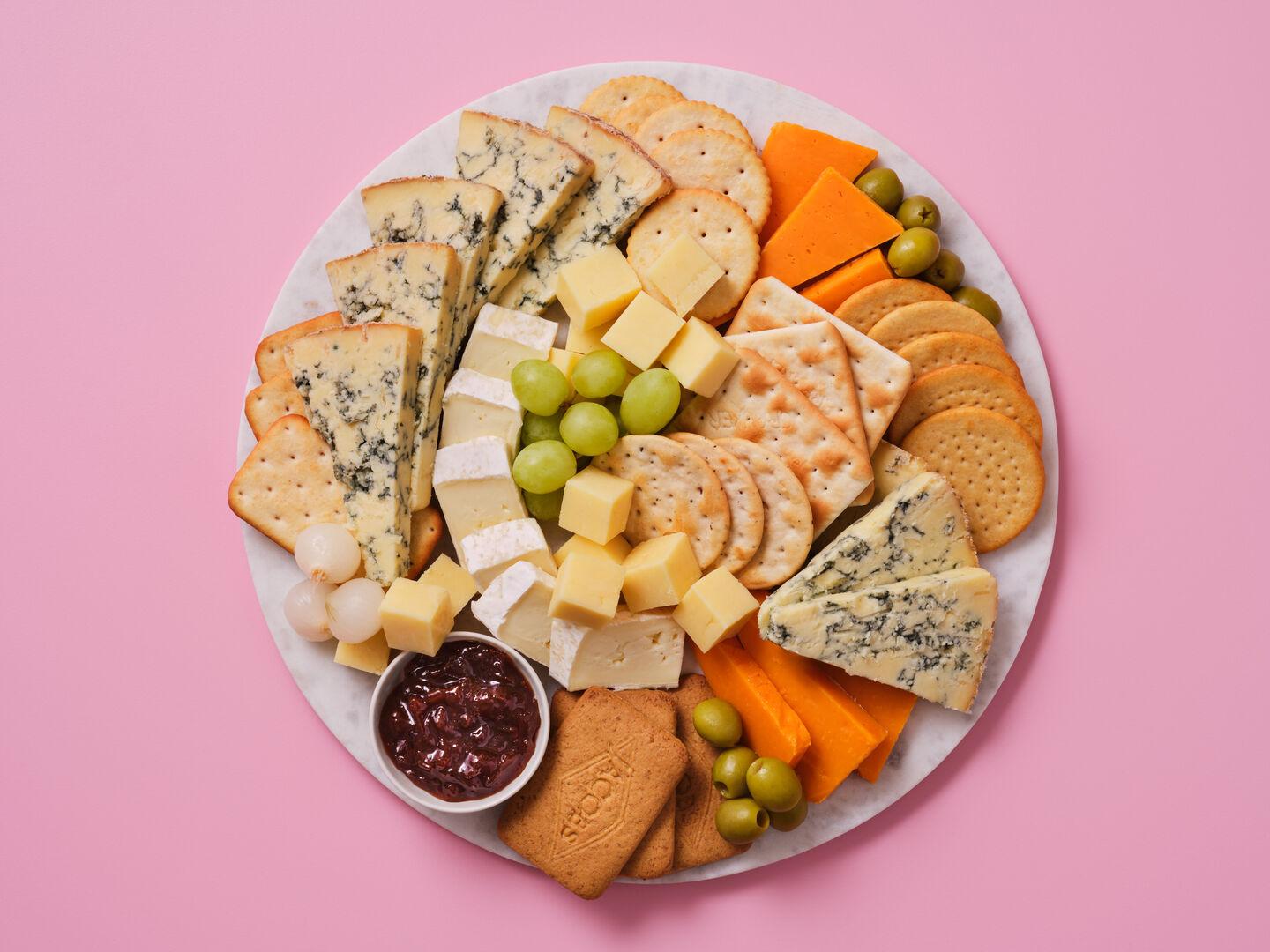 Alt text: A cheeseboard with a selection of blue cheese, brie and red Leicester, served with crackers, biscuits, grapes, olives, pickled onions and chutney, on a pink background.