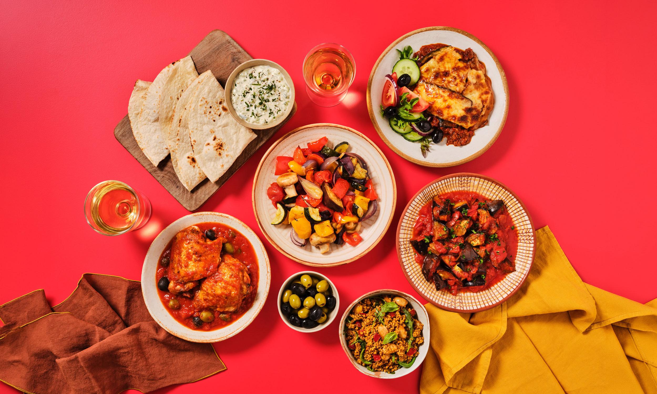 A flatlay of Mediterranean-style dishes arranged on a red background with terracotta and mustard yellow linen napkins. Dishes include chicken in a tomato and olive sauce, a roasted vegetable medley with peppers, courgette, aubergine, and red onion, a moussaka with golden béchamel topping served with a Greek salad of tomato, cucumber, olive, and red onion, a Sicilian-style caponata of aubergine and tomato, and a spiced couscous salad. Accompaniments include flatbreads on a wooden board with a herb yoghurt dip, a bowl of mixed green and black olives, and two glasses of rosé wine.