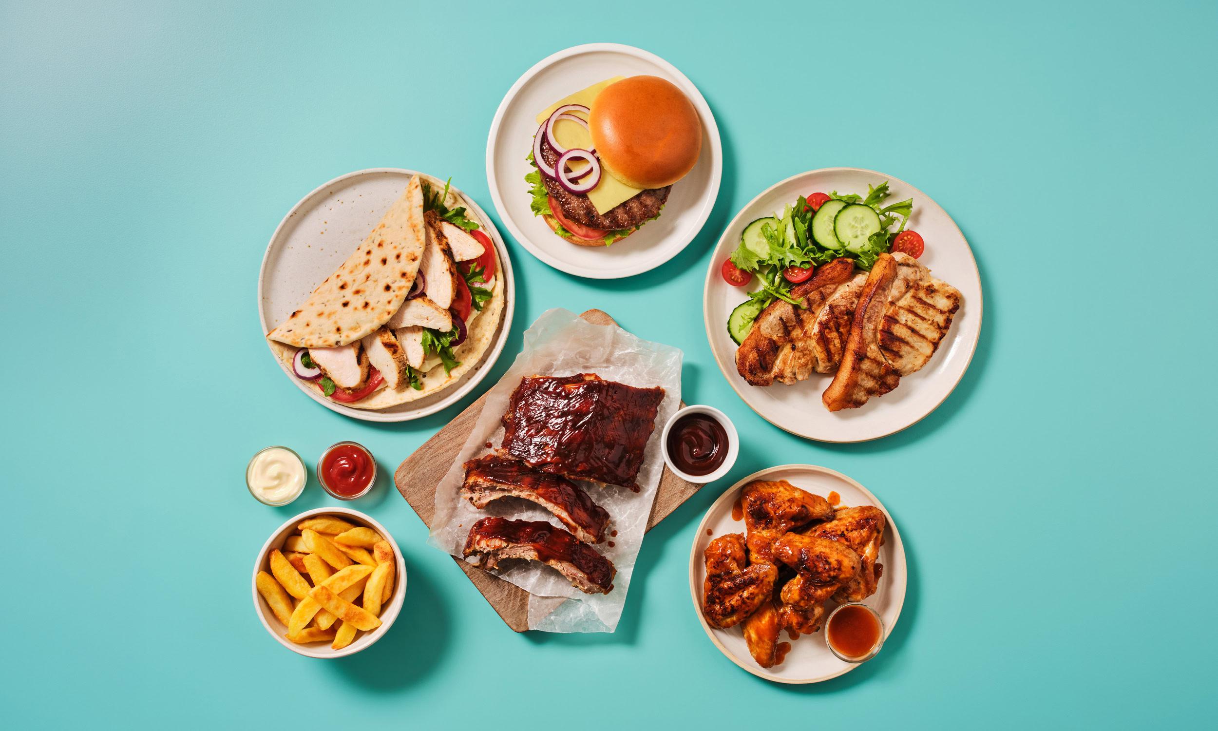 A flatlay of assorted grilled and barbecue dishes arranged on a teal background. Dishes include a chicken wrap with tomato and red onion, a cheeseburger in a brioche bun, grilled chicken with a fresh side salad of lettuce, cucumber, and tomato, sticky barbecue ribs served on a wooden board with parchment paper, and glazed chicken wings with a dipping sauce. A bowl of thick-cut chips, a pot of ketchup, and a pot of mayonnaise are also visible.