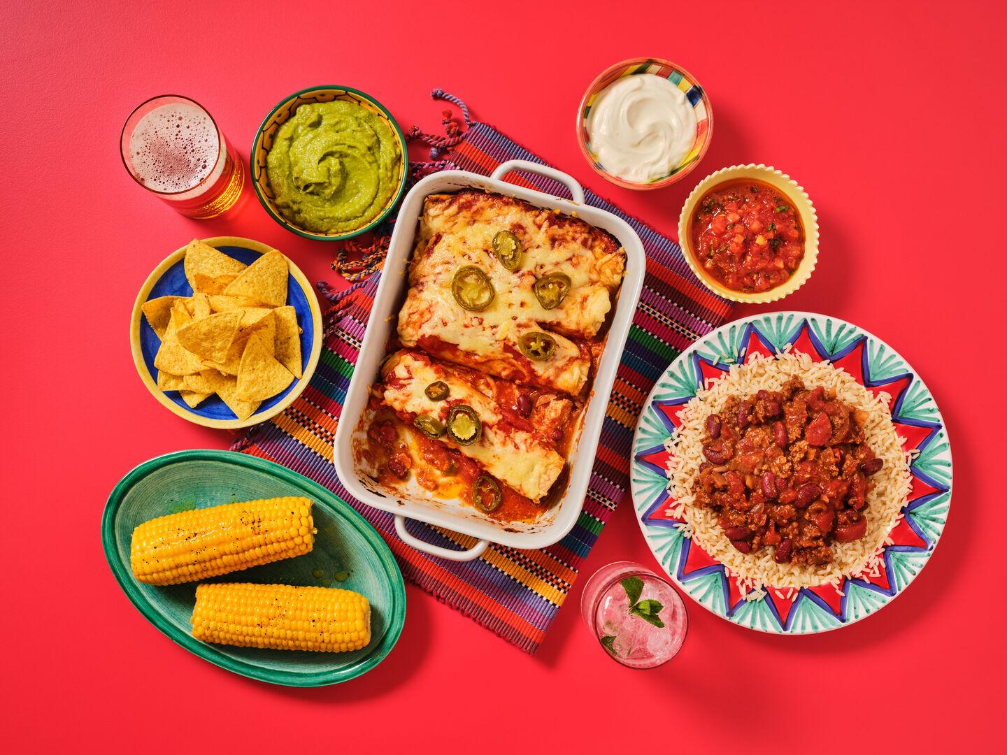 A Mexican-style meal spread on a red background, including cheesy enchiladas, chilli con carne with rice, corn on the cob, tortilla chips, guacamole, salsa and sour cream.