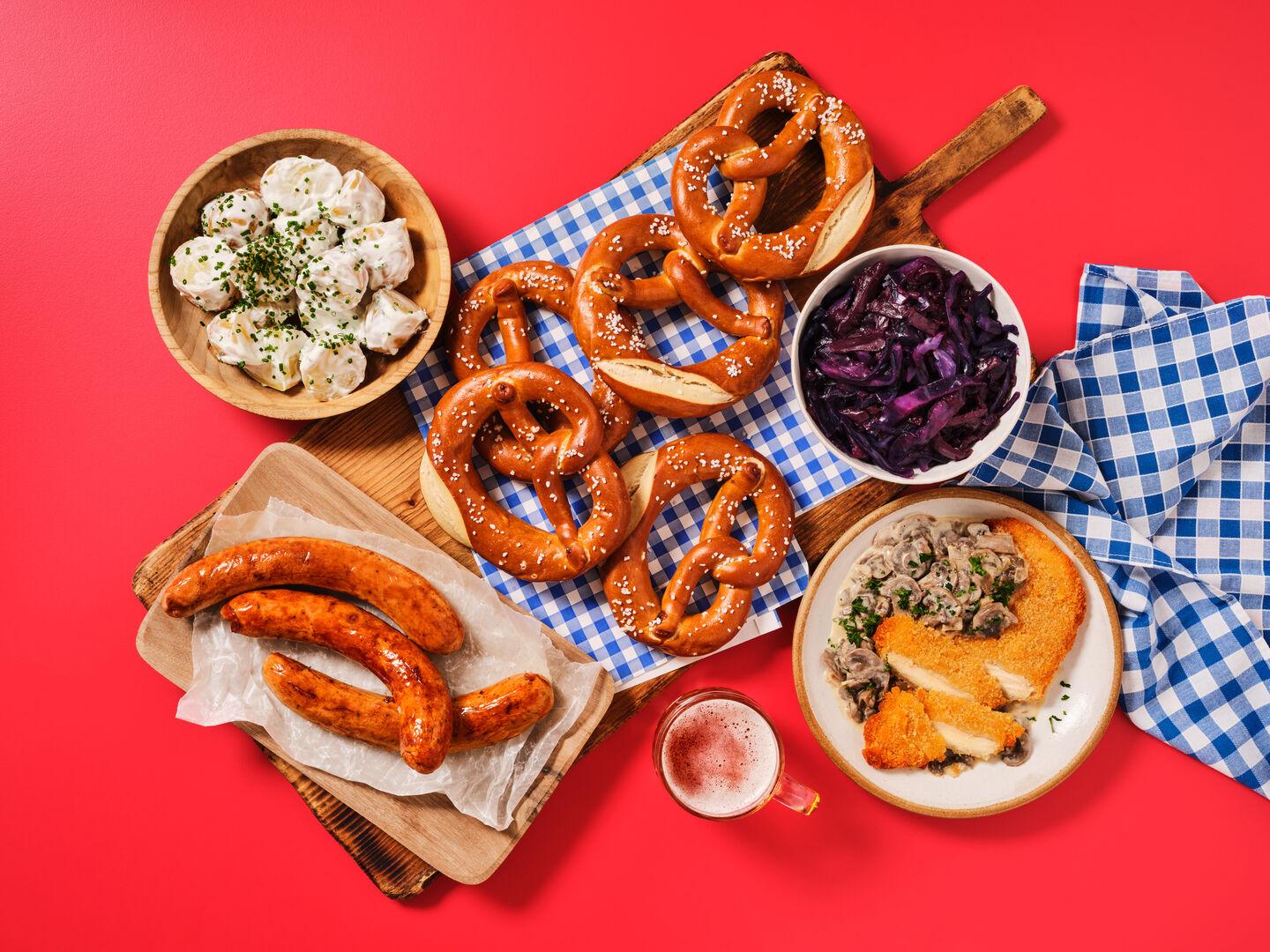 A German-style meal spread on a red background, including pretzels, bratwurst sausages, breaded schnitzel with mushroom sauce, red cabbage and potato salad.