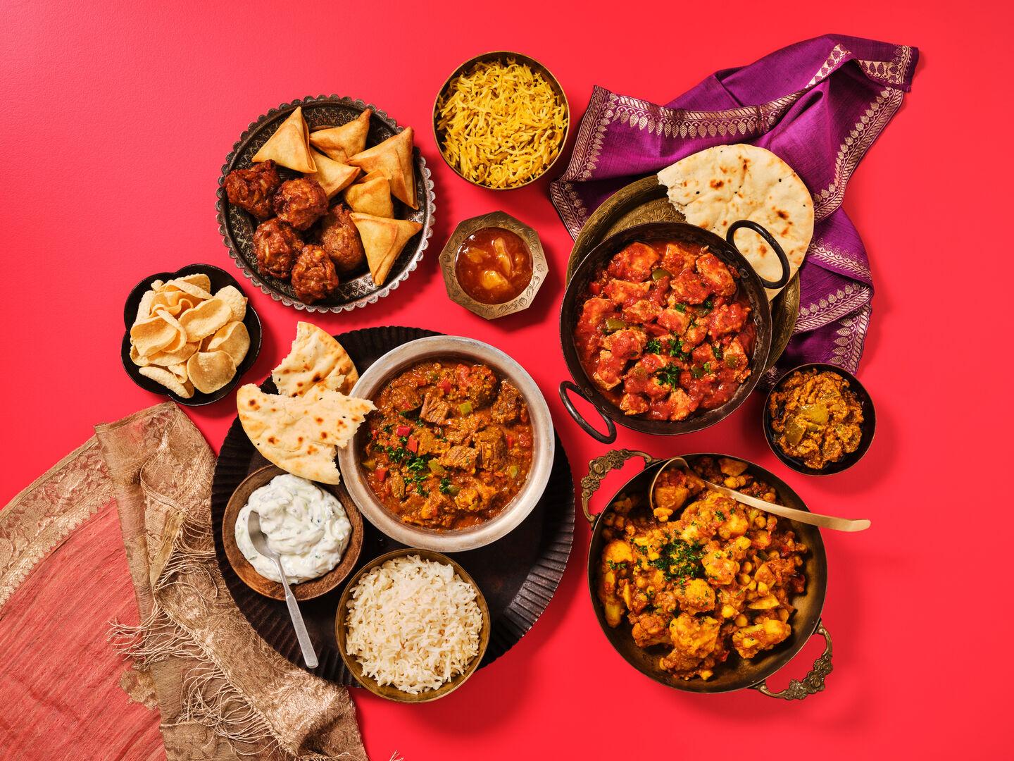 An Indian-style meal spread on a red background, including various curries, rice, naan bread, poppadoms, samosas, onion bhajis, raita and chutney.