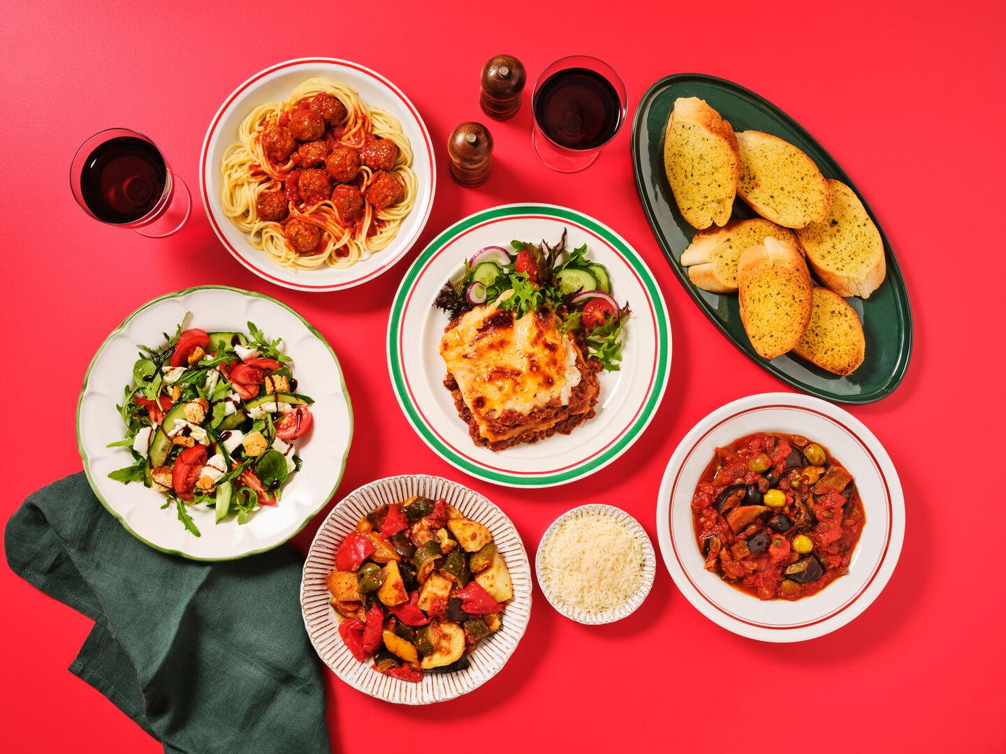 An Italian-style meal spread on a red background, including spaghetti and meatballs, lasagne, garlic bread, roasted vegetables, aubergine caponata, salad and glasses of red wine.