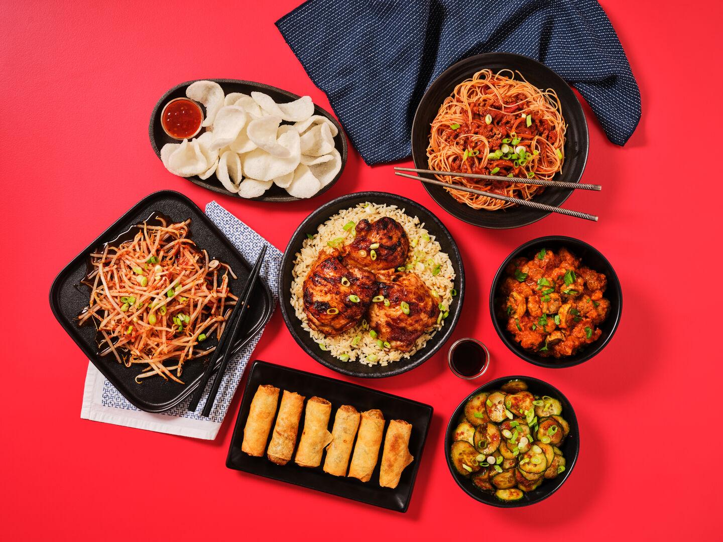 A spread of Asian-inspired dishes on a red background, including chicken and rice, chow mein, spring rolls, prawn crackers, bean sprouts, kung po and stir-fried courgette.
