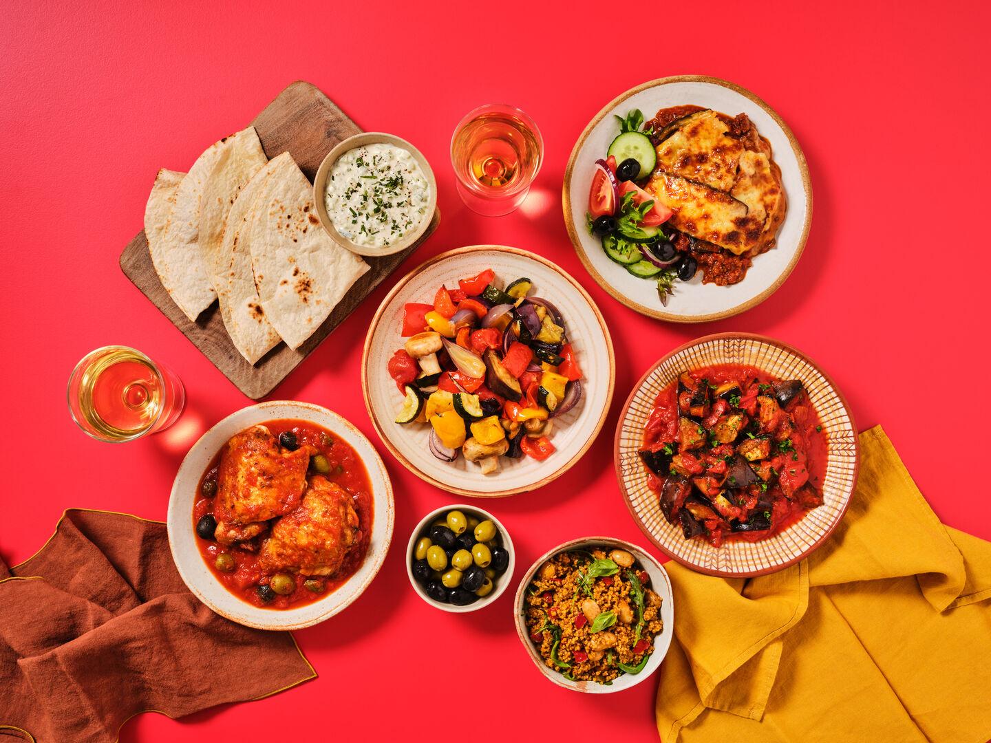 A spread of Mediterranean dishes on a red background, including moussaka, chicken cacciatore, ratatouille, flatbreads with tzatziki, olives and a grain salad, with glasses of rosé wine.