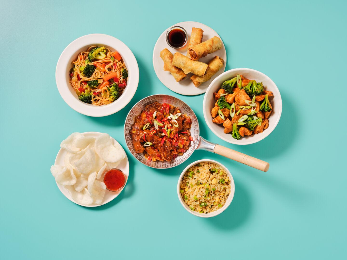 A Chinese-style meal spread on a teal background, including chow mein, spring rolls, sweet and sour chicken, chicken and broccoli stir fry, egg fried rice and prawn crackers.