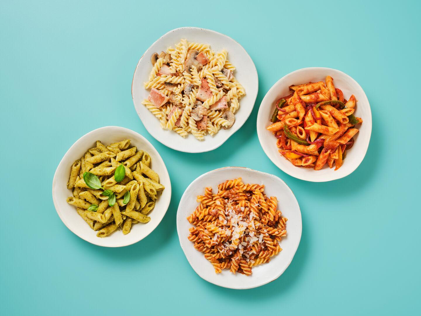 Four bowls of pasta on a teal background, including creamy bacon and mushroom fusilli, pesto penne with basil, spicy penne arrabbiata with peppers, and fusilli bolognese topped with parmesan.