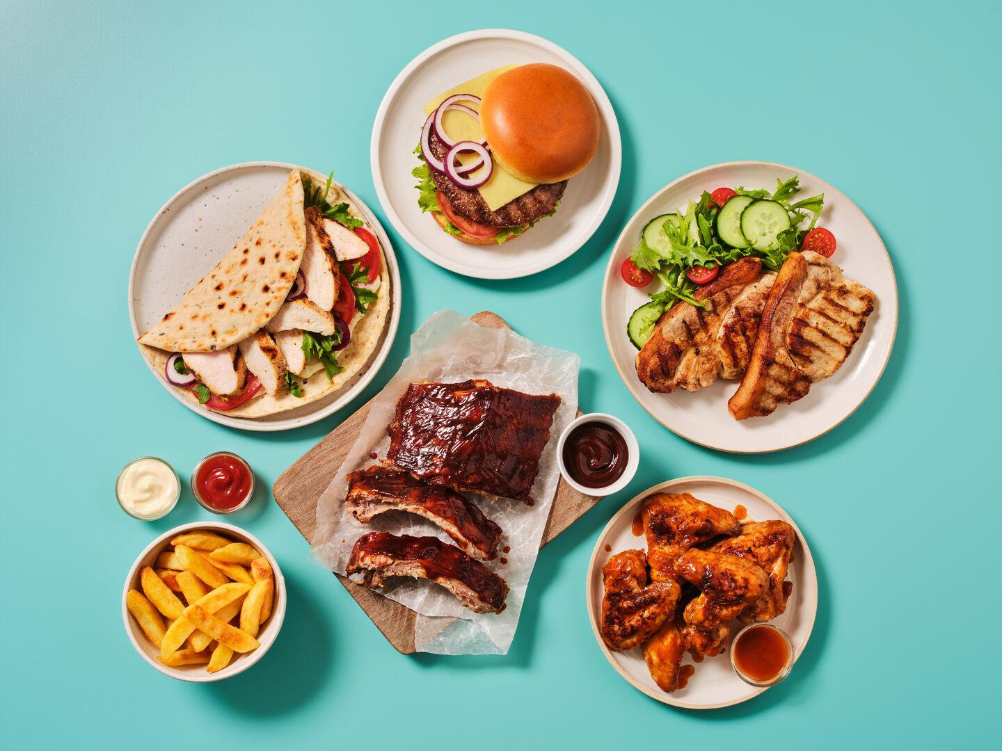 A selection of grilled dishes on a teal background, including a cheeseburger, BBQ ribs, chicken wings, grilled pork chop with salad, a chicken flatbread wrap and a bowl of chips.