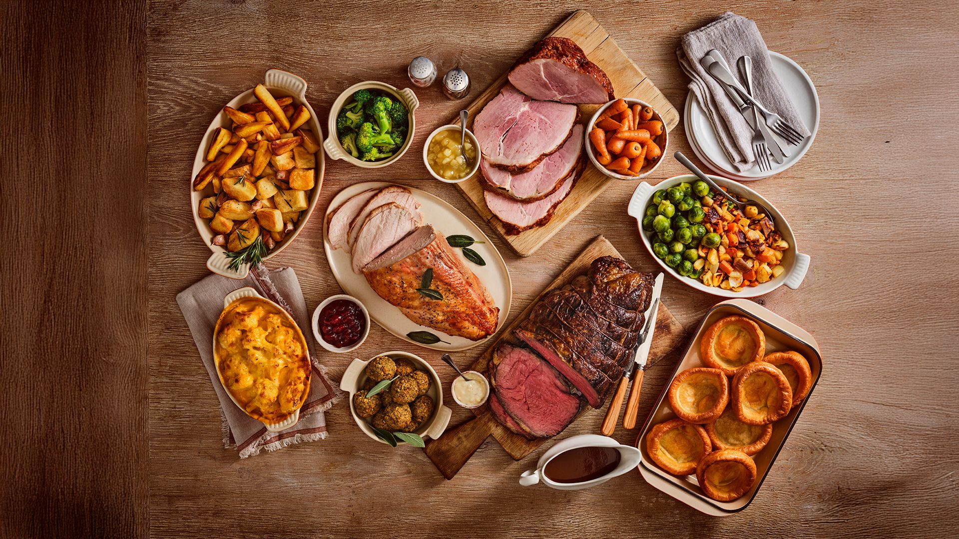 A selection of roast dishes sit on a wooden table. There's a selection of meets, Yorkshire puddings and vegetables.