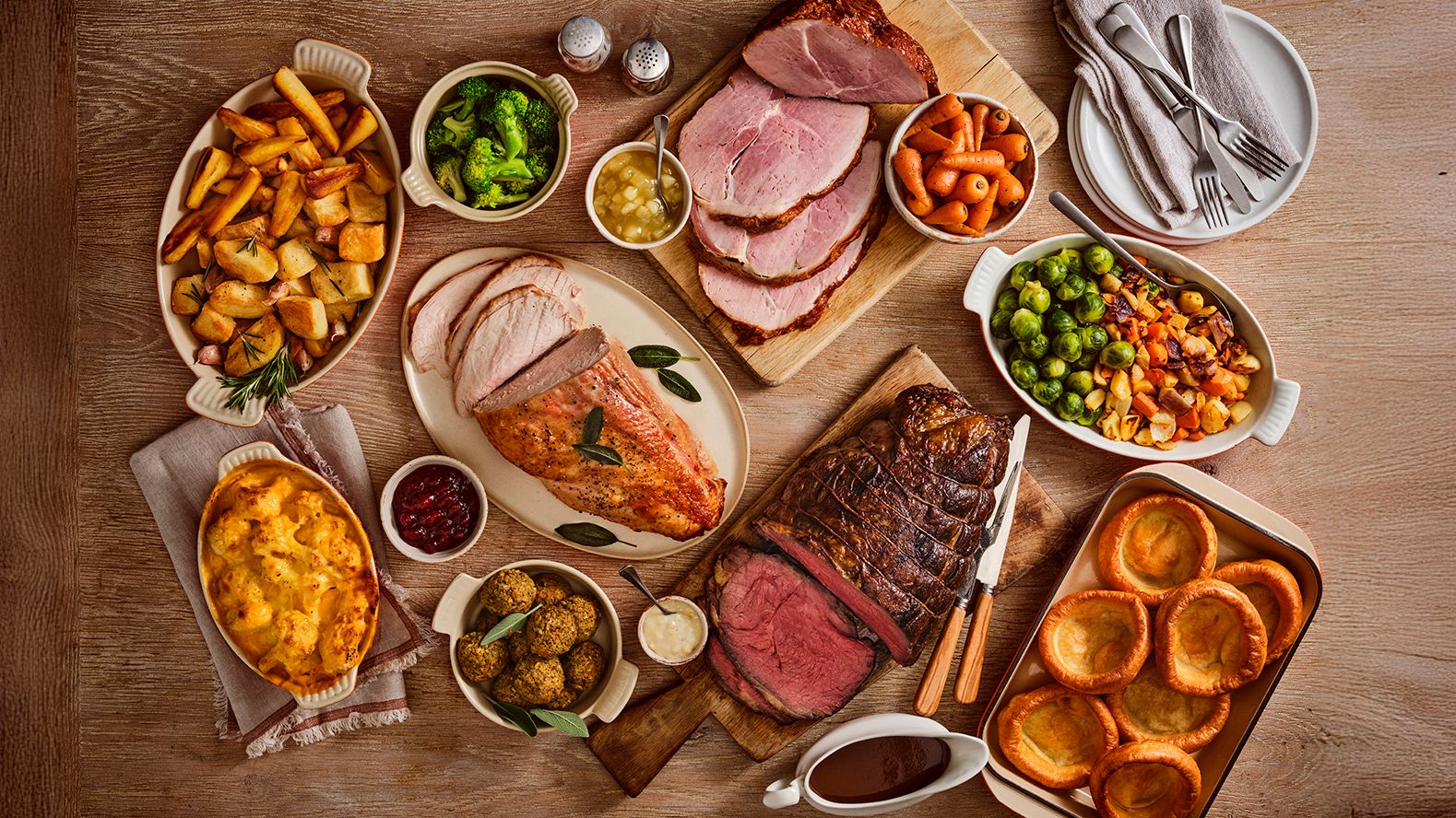 A selection of roast dishes sit on a wooden table. There's a selection of meets, Yorkshire puddings and vegetables.