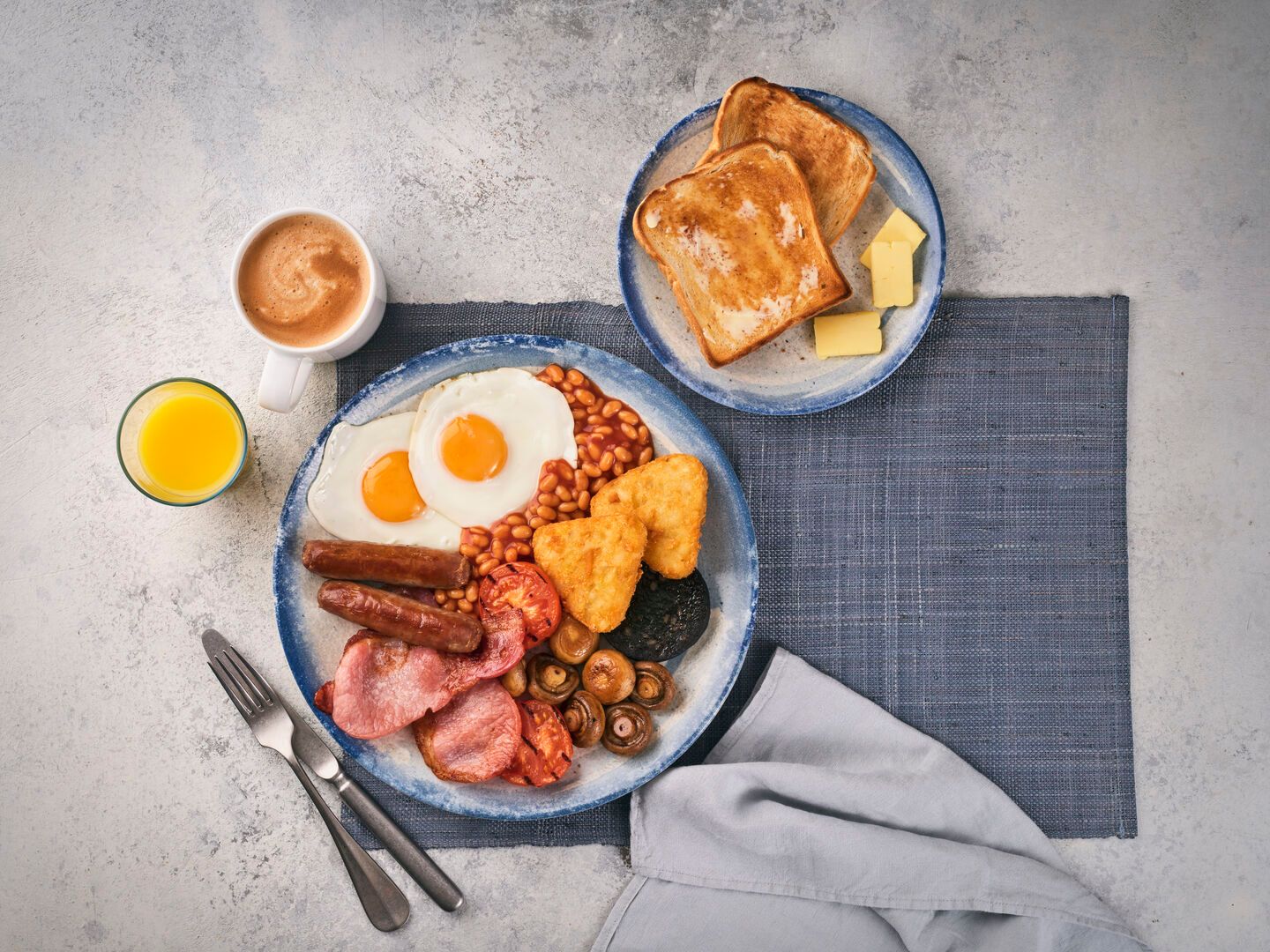 A photo of a full English breakfast, with a plate of two slices of toast to the side alongside a cup of coffee and glass of orange juice.