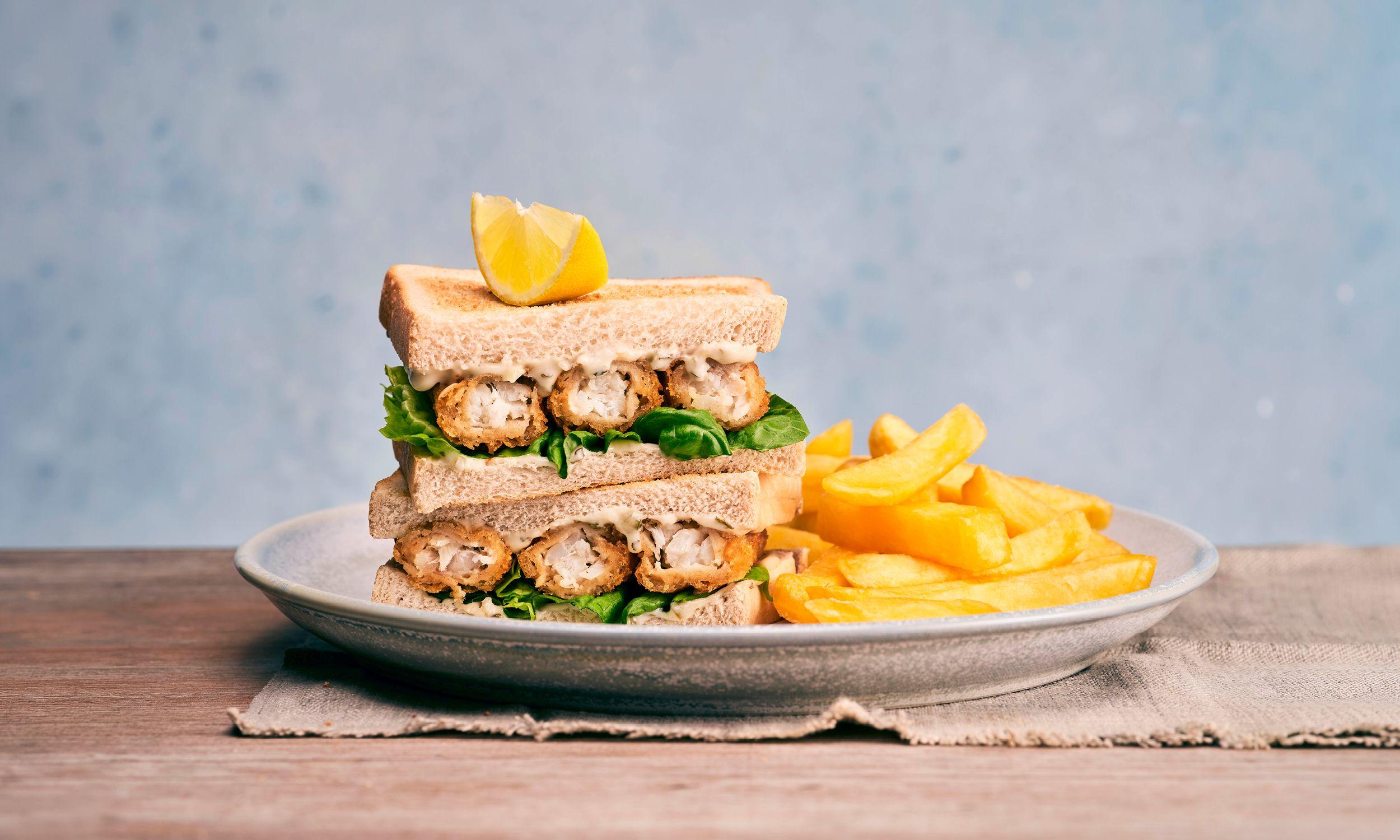 Stacked fish finger sandwich with lettuce and tartare sauce, garnished with a lemon wedge, served with chips on a plate.