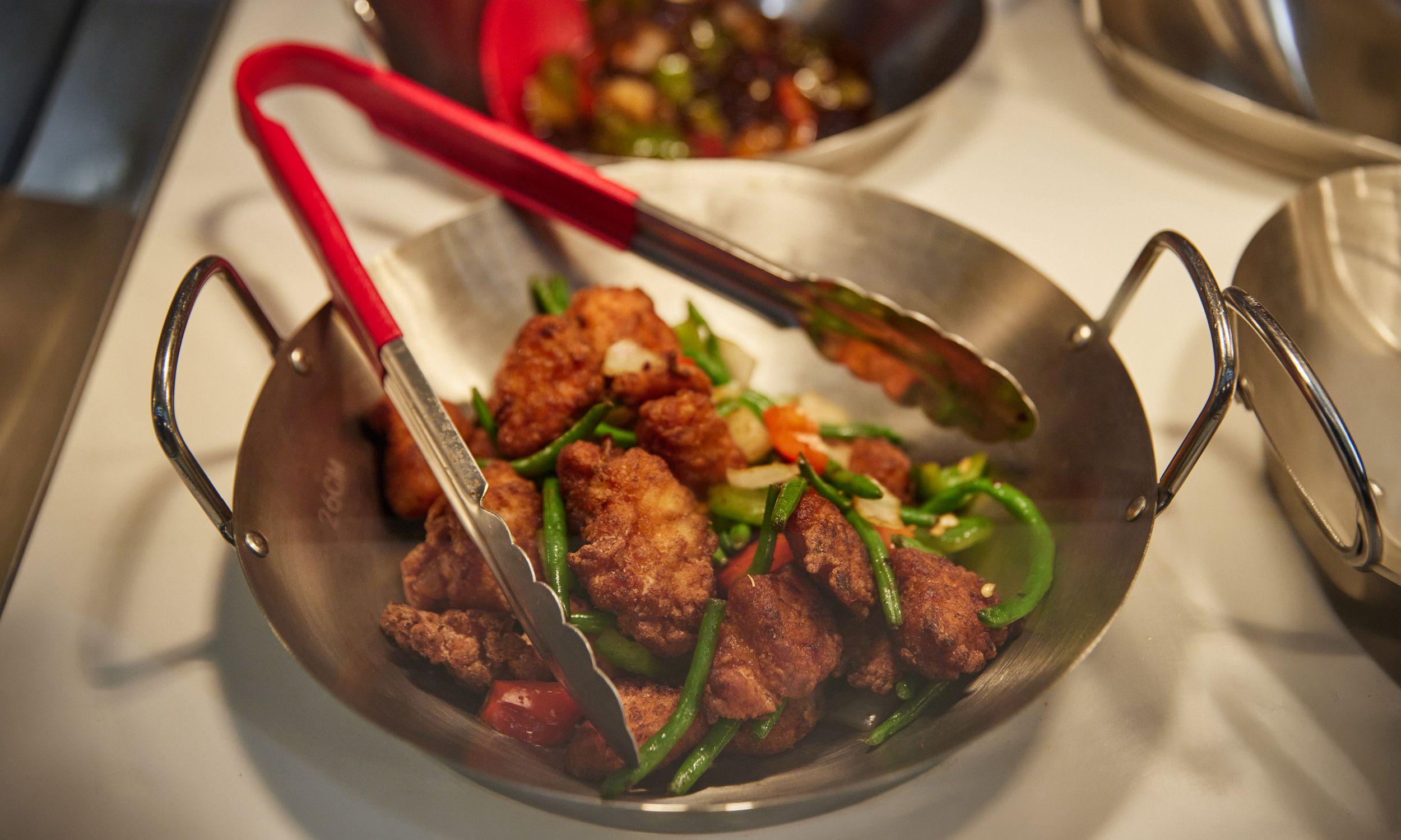 Close-up of crispy fried chicken pieces with green beans and peppers in a metal serving dish, with tongs resting on top.