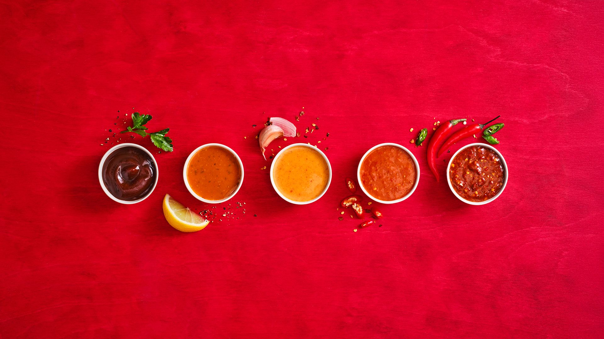 Five different condiments are placed in line on a red table. Between them are different garnishes including parsley, garlic and chilli peppers.