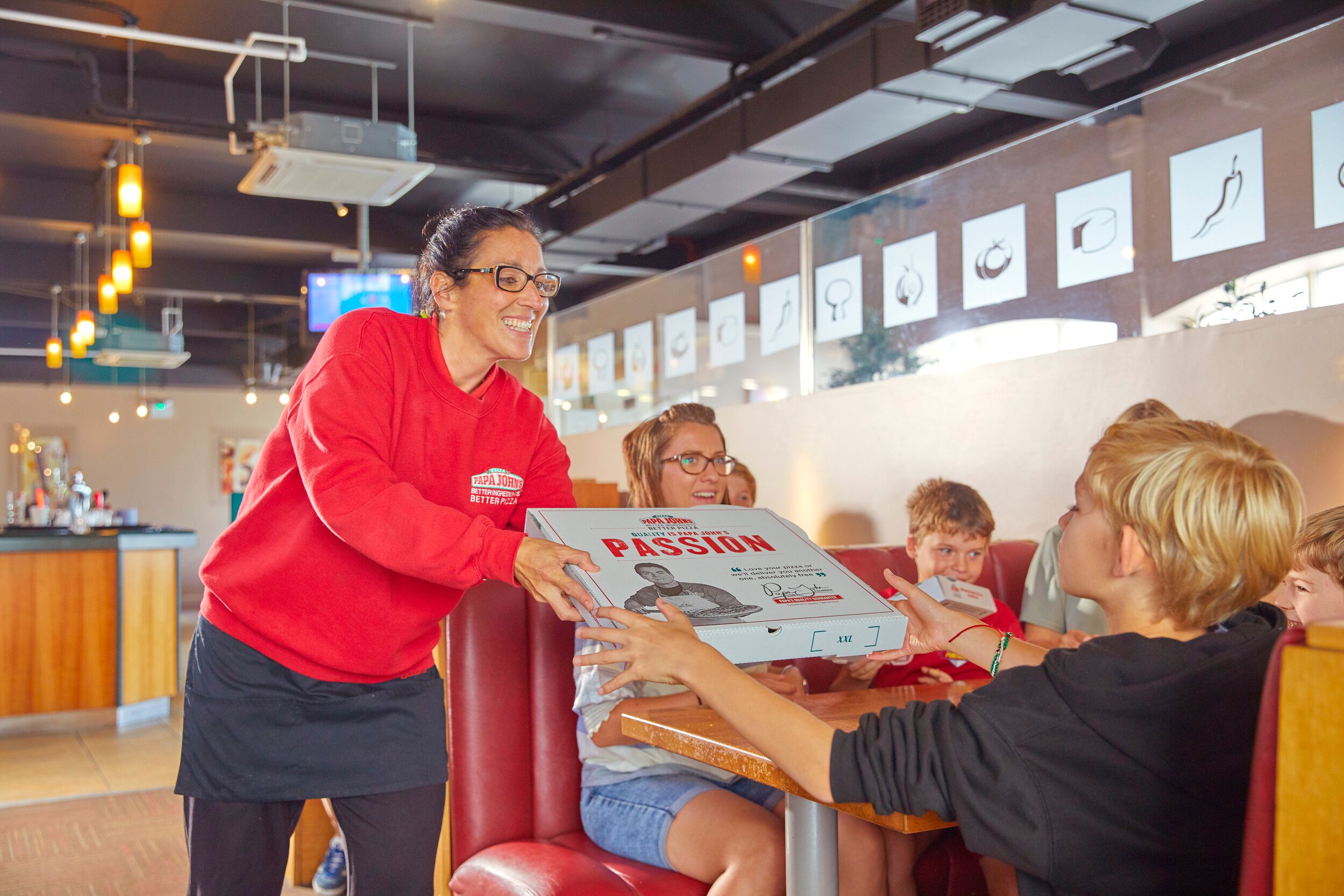 A staff of Papa John's handing a box of pizza to a family around the dining table.
