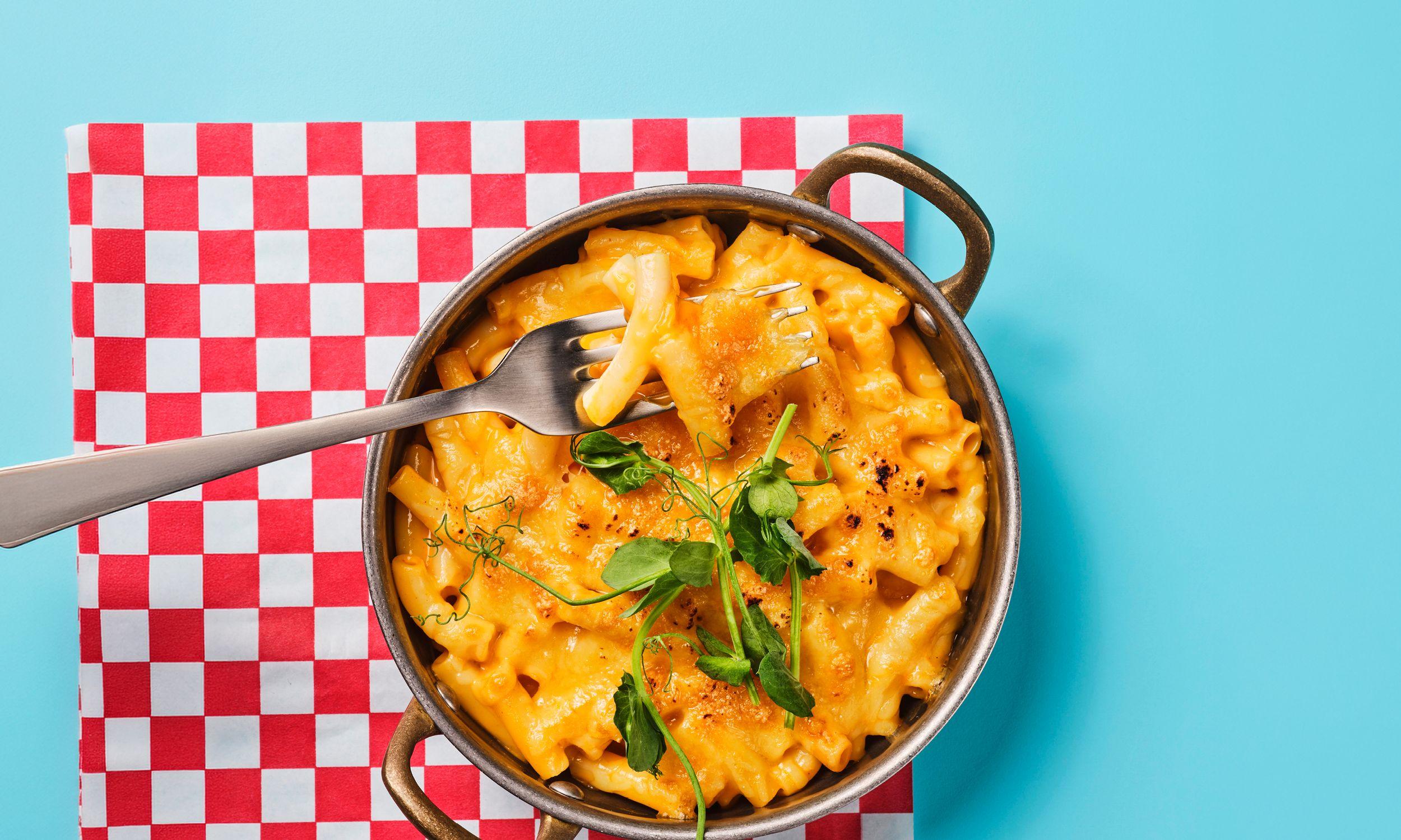 Overhead shot of a baked mac and cheese in a metal pot, garnished with pea shoots, with a fork lifting a bite, set on a red-and-white checked napkin.