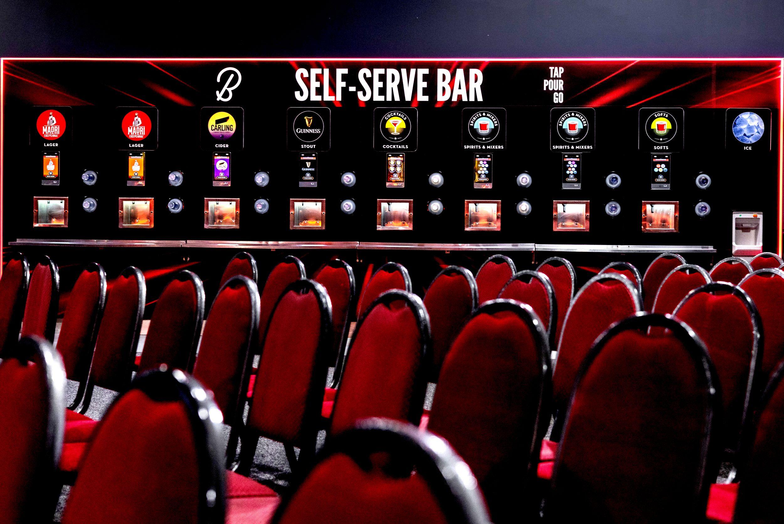 Boxbar self-serve bar wall with 'Tap Pour Go' signage, featuring touchscreen dispensers for Madrí lager (x2), Carling cider, Guinness stout, cocktails, spirits and mixers (x2), softs, and ice, viewed from behind rows of red venue seating.