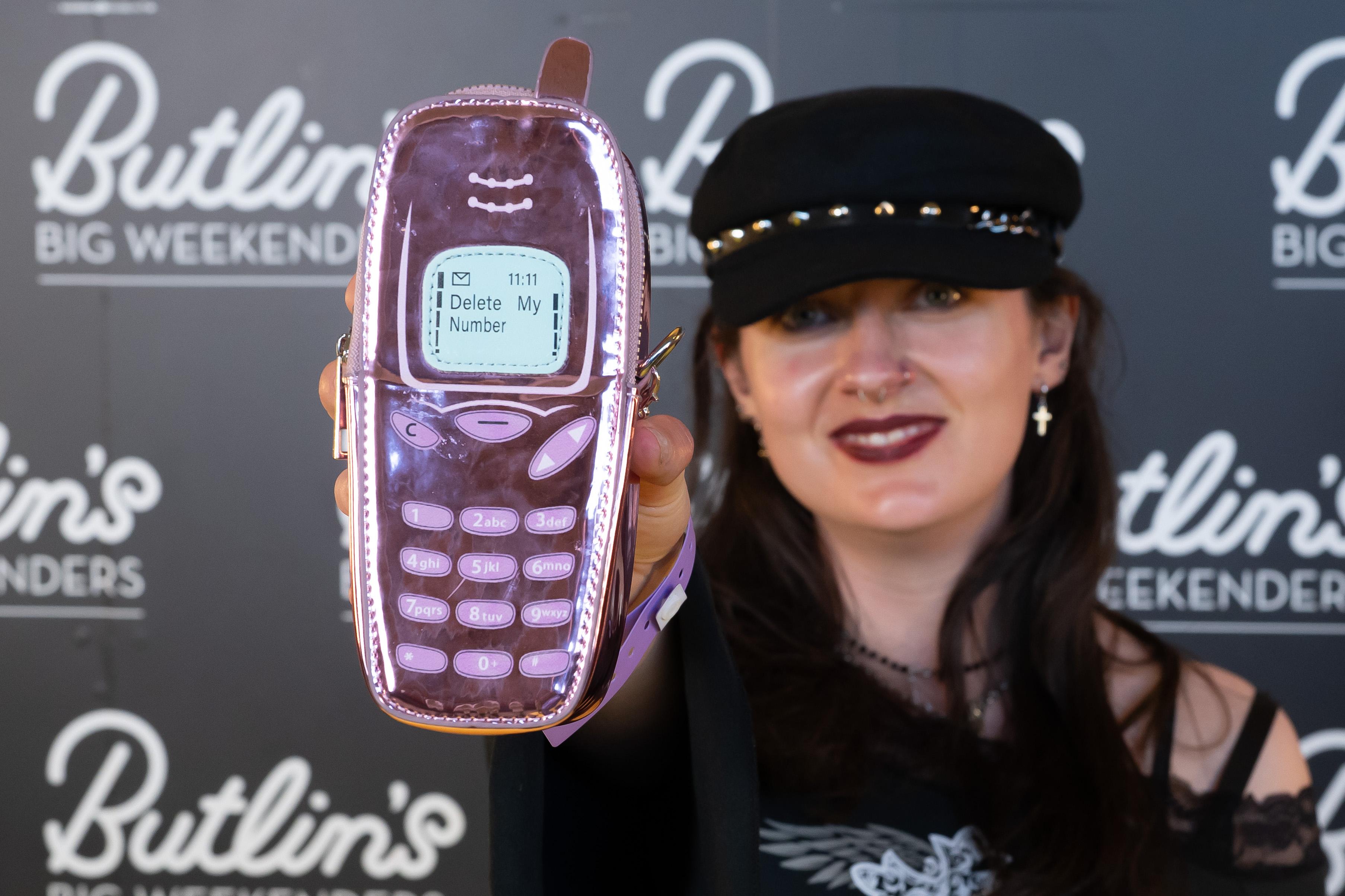 A person holds up a pink metallic Nokia-style phone-shaped bag displaying the message 'Delete My Number' on its screen, in front of a Butlin's Big Weekenders branded backdrop.