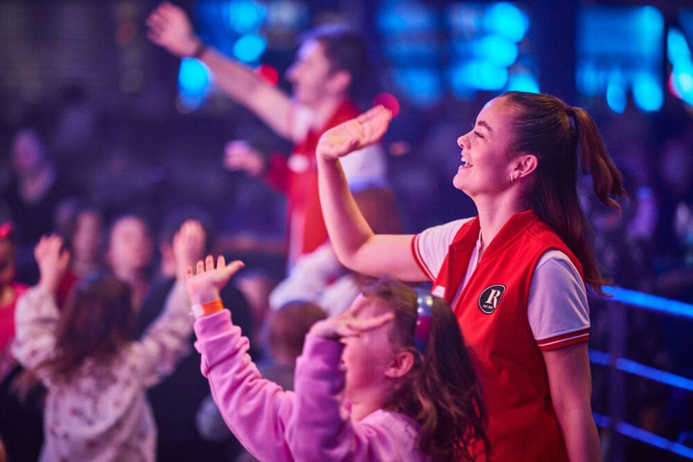 A Redcoat waves towards the stage while children around her do the same.