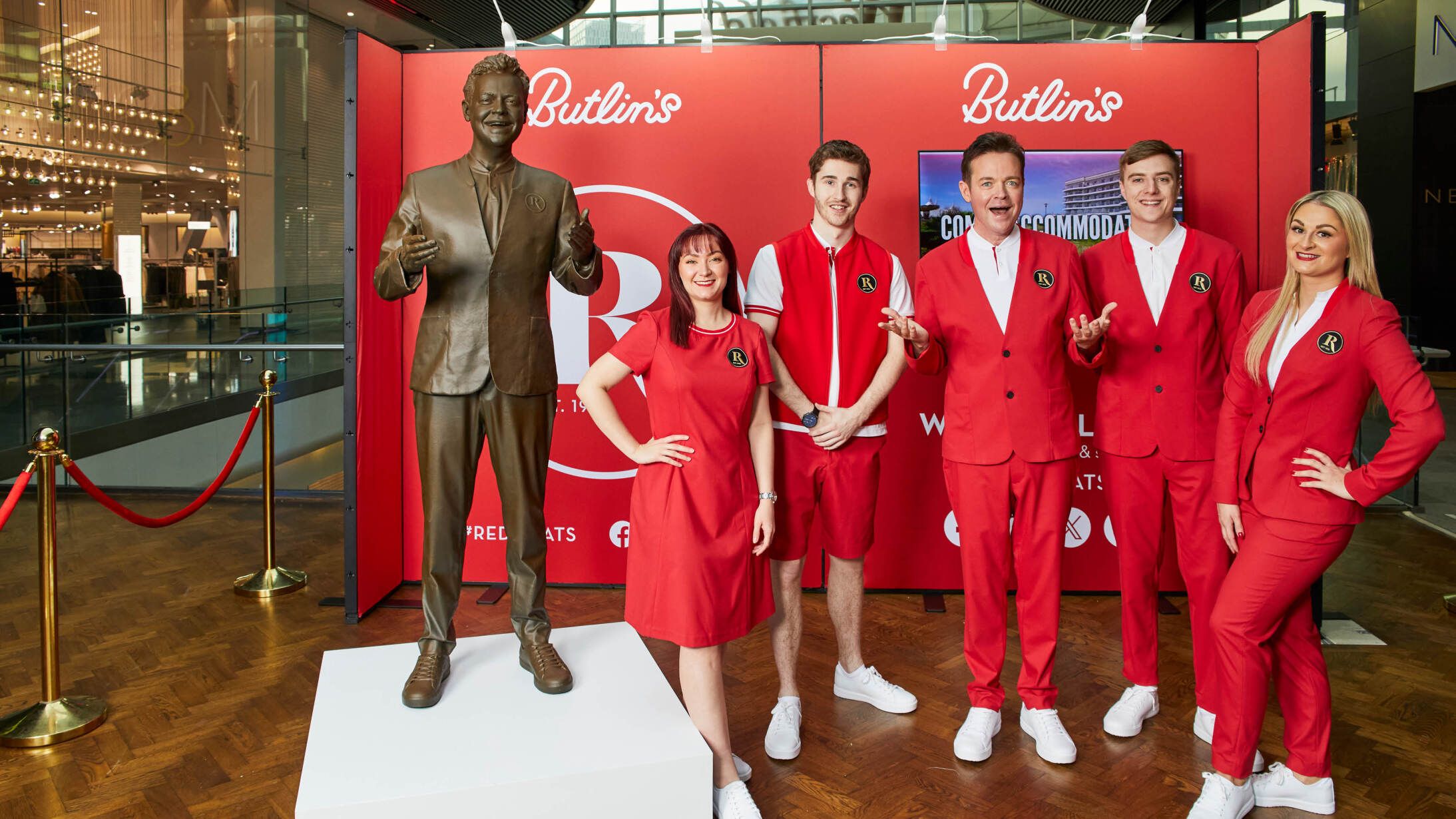 Stephen Mulhern wears a Redcoat uniform, and stands alongside other Redcoat team members. To their left is a statue of Stephen Mulhern.