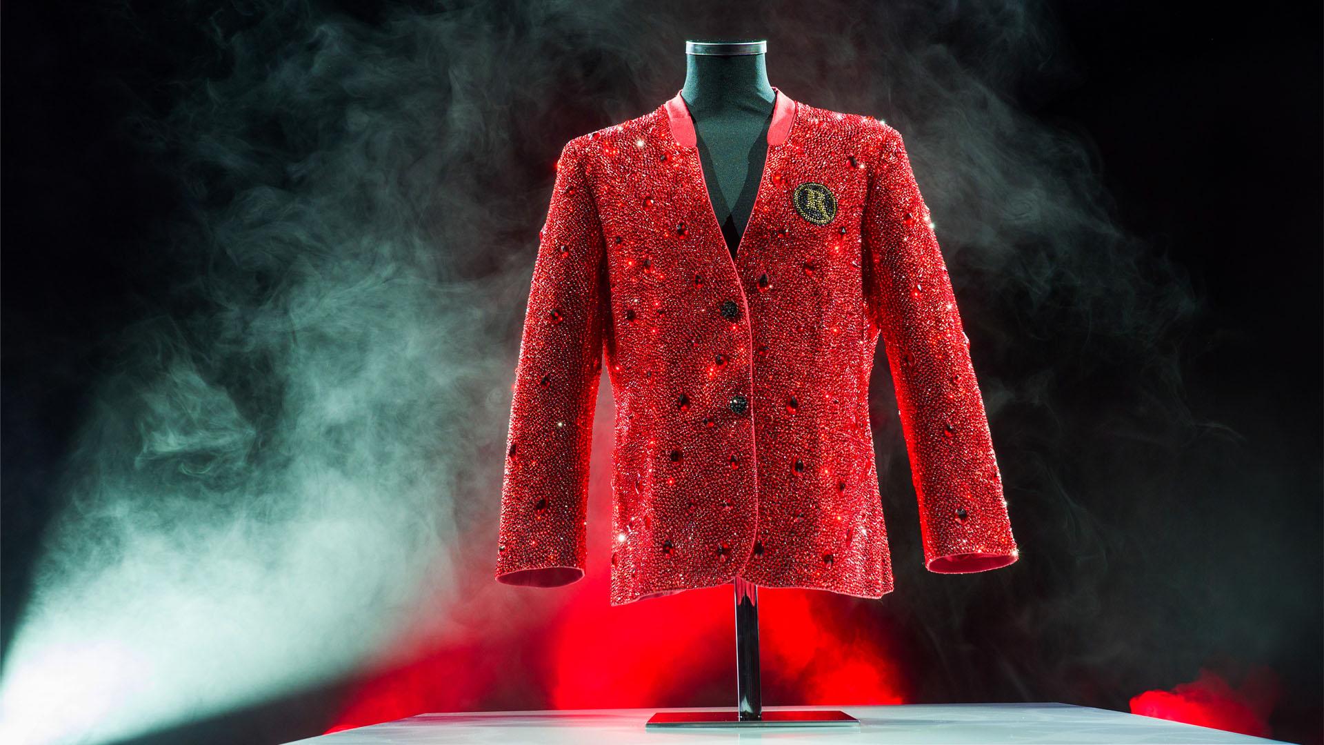A red crystal-encrusted blazer covered in thousands of red rhinestones and scattered larger gemstones, displayed on a black mannequin torso against a dramatic dark background with swirling smoke and red lighting. A circular embroidered crest with the letter 'R' is visible on the left chest.