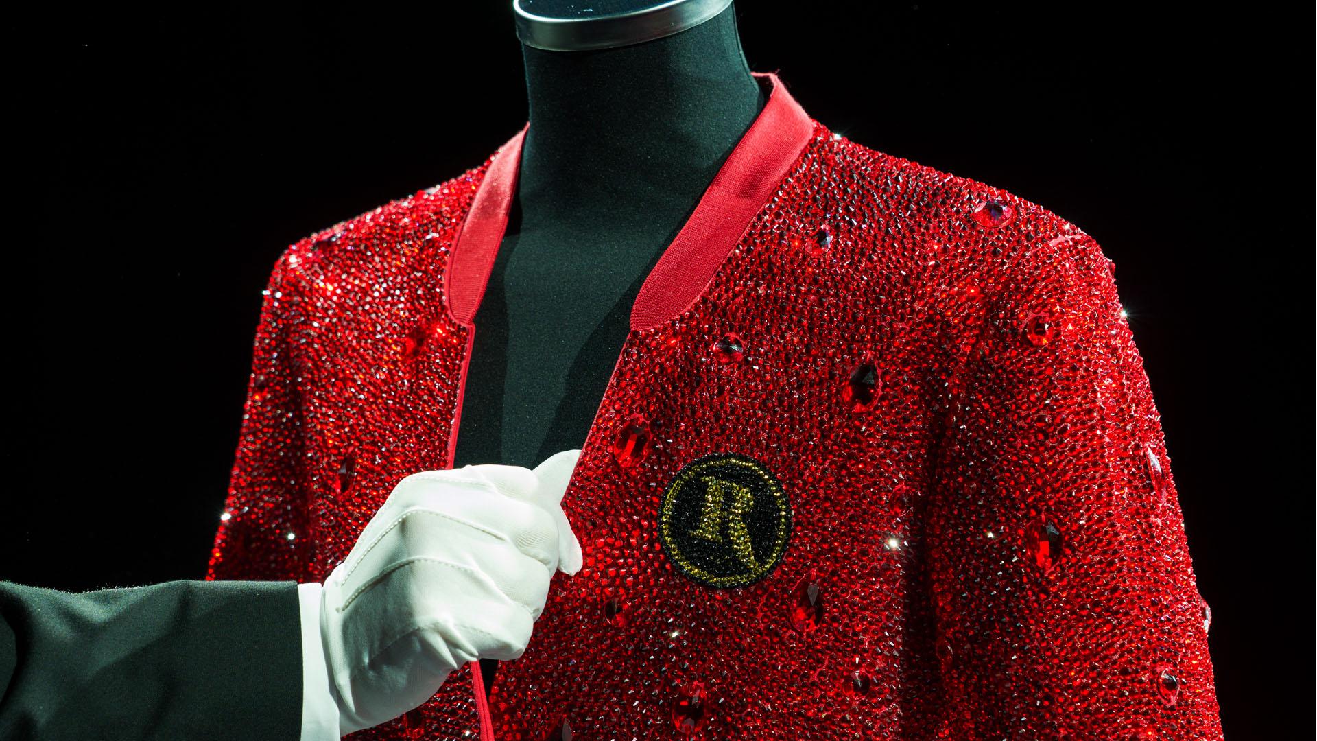 Close-up of a red rhinestone-covered blazer on a mannequin, with a white-gloved hand pointing to a circular beaded crest on the chest. The crest features a gold letter 'R' on a black background, surrounded by intricate gold beadwork.