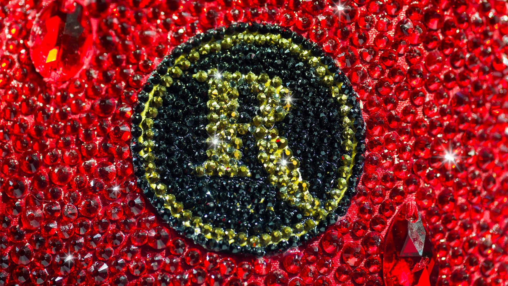 Extreme macro close-up of a circular crystal emblem on a red rhinestone garment. The badge features a stylized letter 'R' rendered in gold/yellow crystals set against a background of black crystals, surrounded by densely packed red faceted rhinestones.